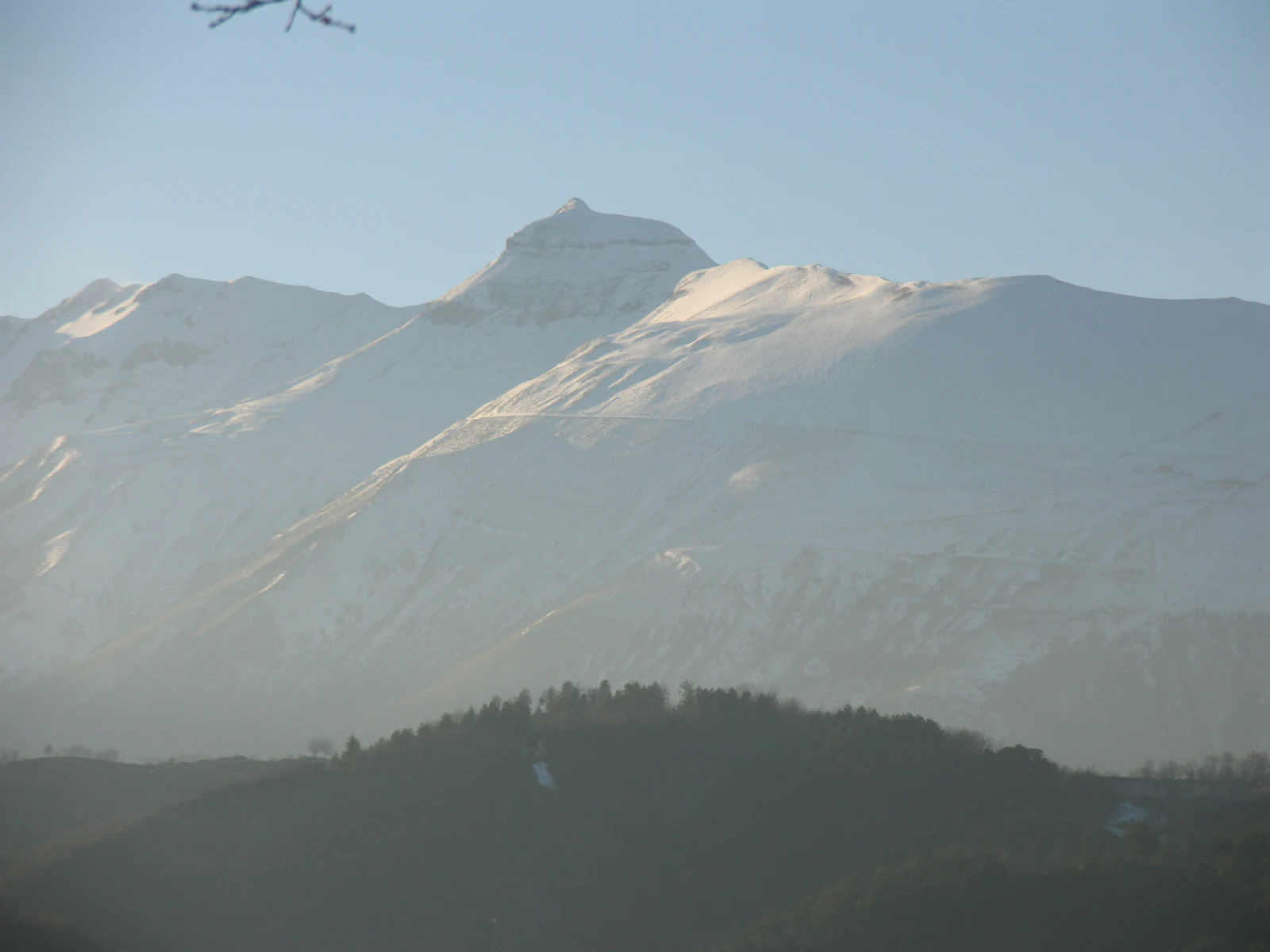 Snow-capped mountain peak with forested hills in the foreground under a clear sky.