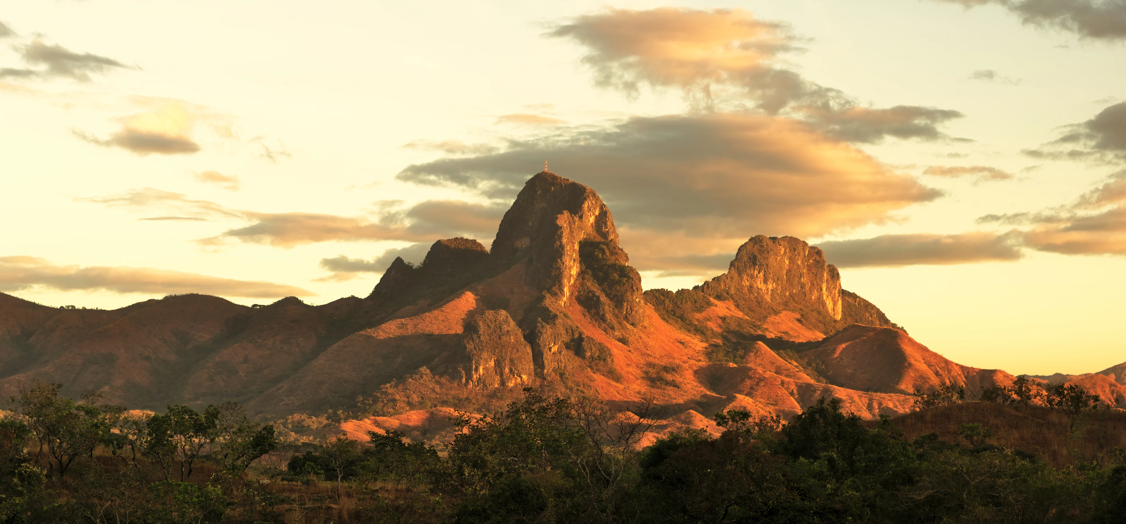 A landscape view of several towering, craggy limestone rock formations, known as the Morros de San Juan, glowing in the warm golden light of sunset against a soft, partly cloudy sky.