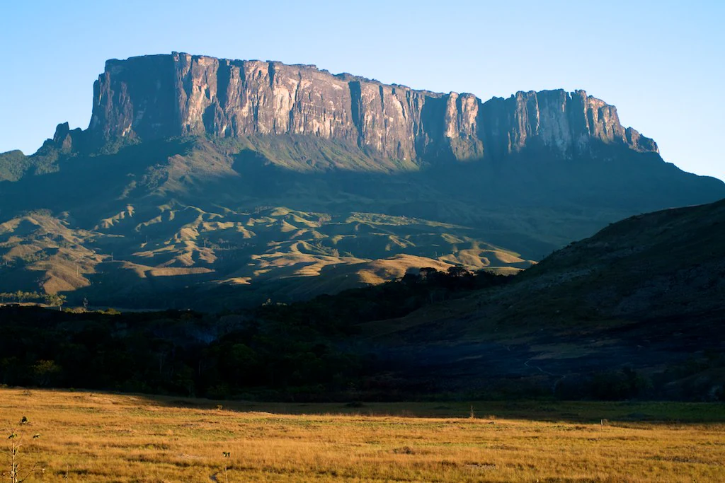Wide view of a large, flat-topped tepui mountain rising above a grassy savanna at sunset.