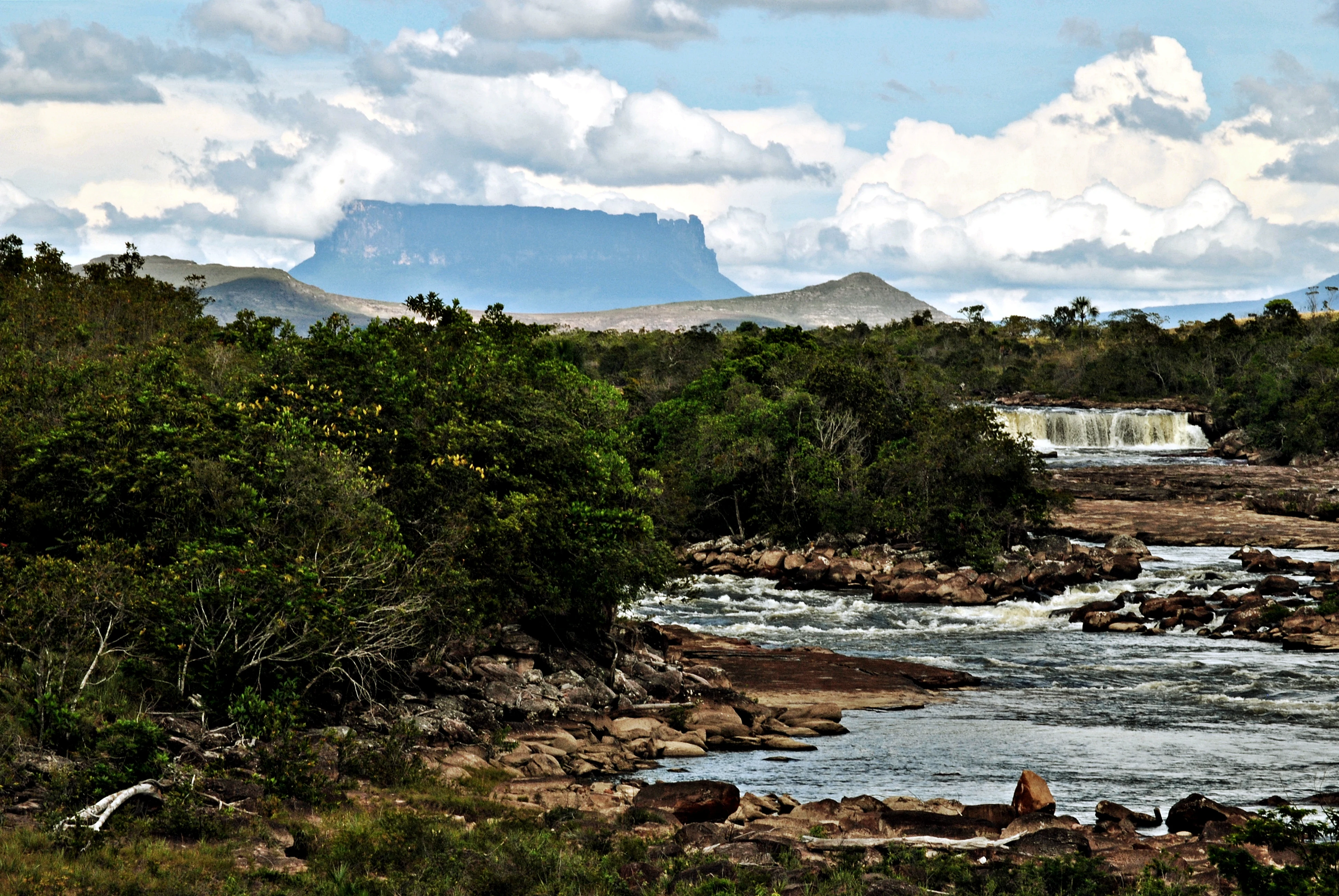 A wide landscape view of a river flowing over rocks toward a small waterfall, with a flat-topped tepuy mountain rising in the distant background under a cloudy sky.