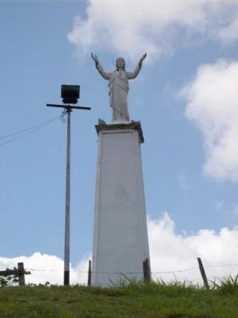 A white statue of Christ with outstretched arms standing on a tall, rectangular concrete pedestal, set against a bright blue sky with scattered clouds.