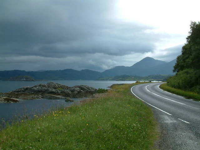 A winding asphalt road curves alongside a rocky shoreline and calm water, with rolling green hills and mountains under a cloudy sky.