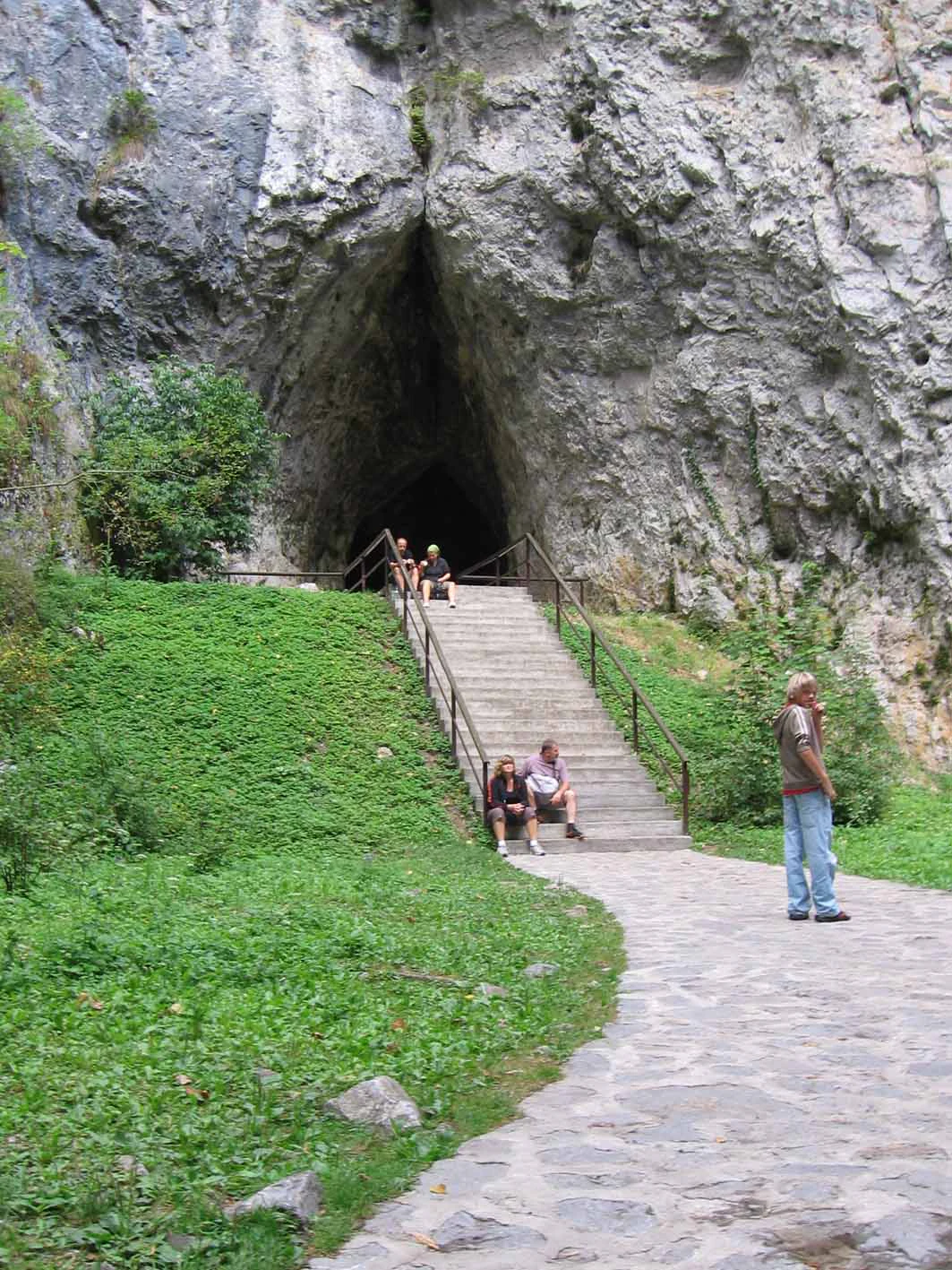 Stone staircase leading to cave entrance in rocky cliff, with visitors on steps and green vegetation