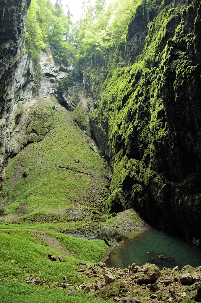 Deep gorge with steep moss-covered cliffs, green slopes, and a small body of water at the base