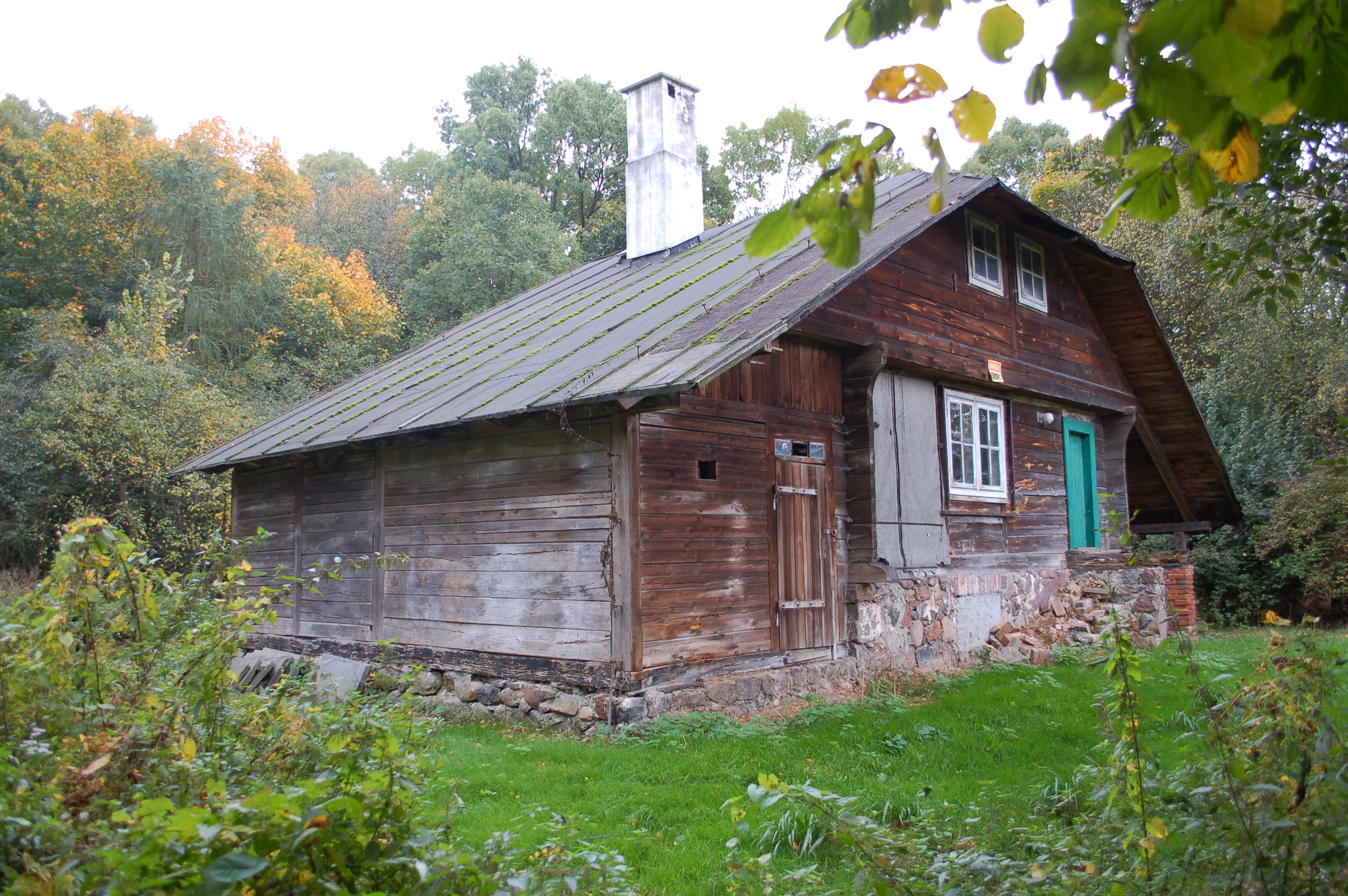 A rustic wooden cabin with a tall white chimney, situated in a forested area.