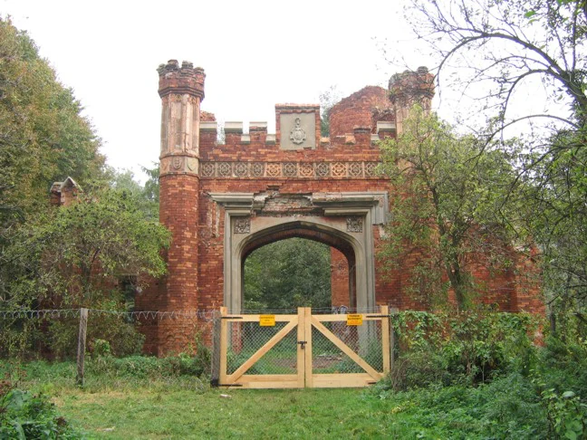 An old, red brick ruins of a gatehouse with crenellated towers and a stone arch, featuring a new wooden gate in the foreground, set against a backdrop of trees.