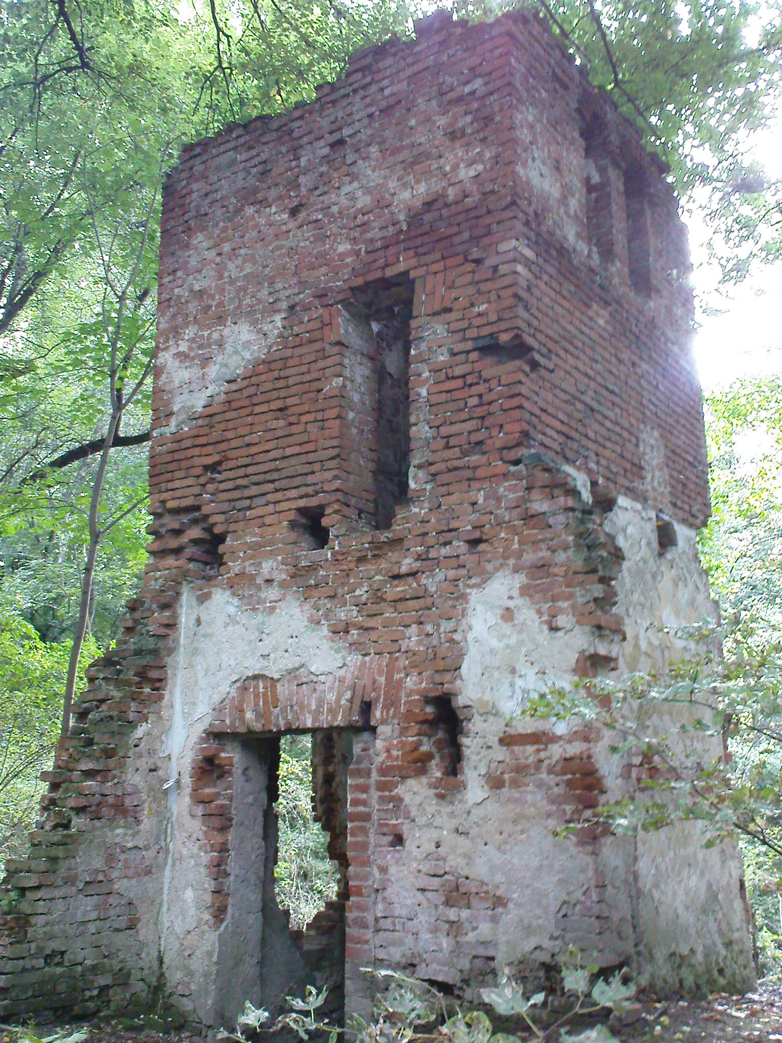 A crumbling two-story red brick ruin, formerly a gatekeeper's house, standing in a wooded area with exposed masonry and partial plaster.