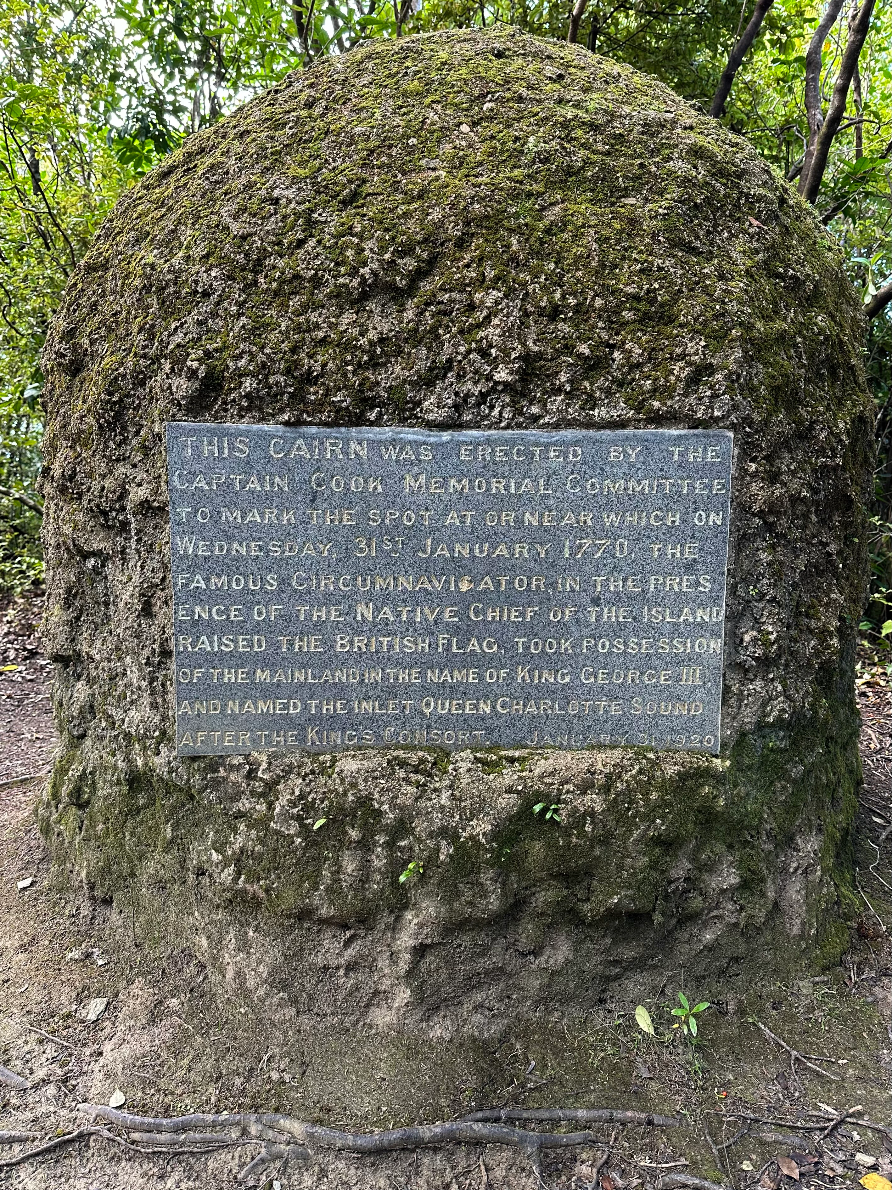 A weathered stone cairn covered in moss with an embedded metal plaque describing the 1770 historical event involving Captain James Cook on Motuara Island.