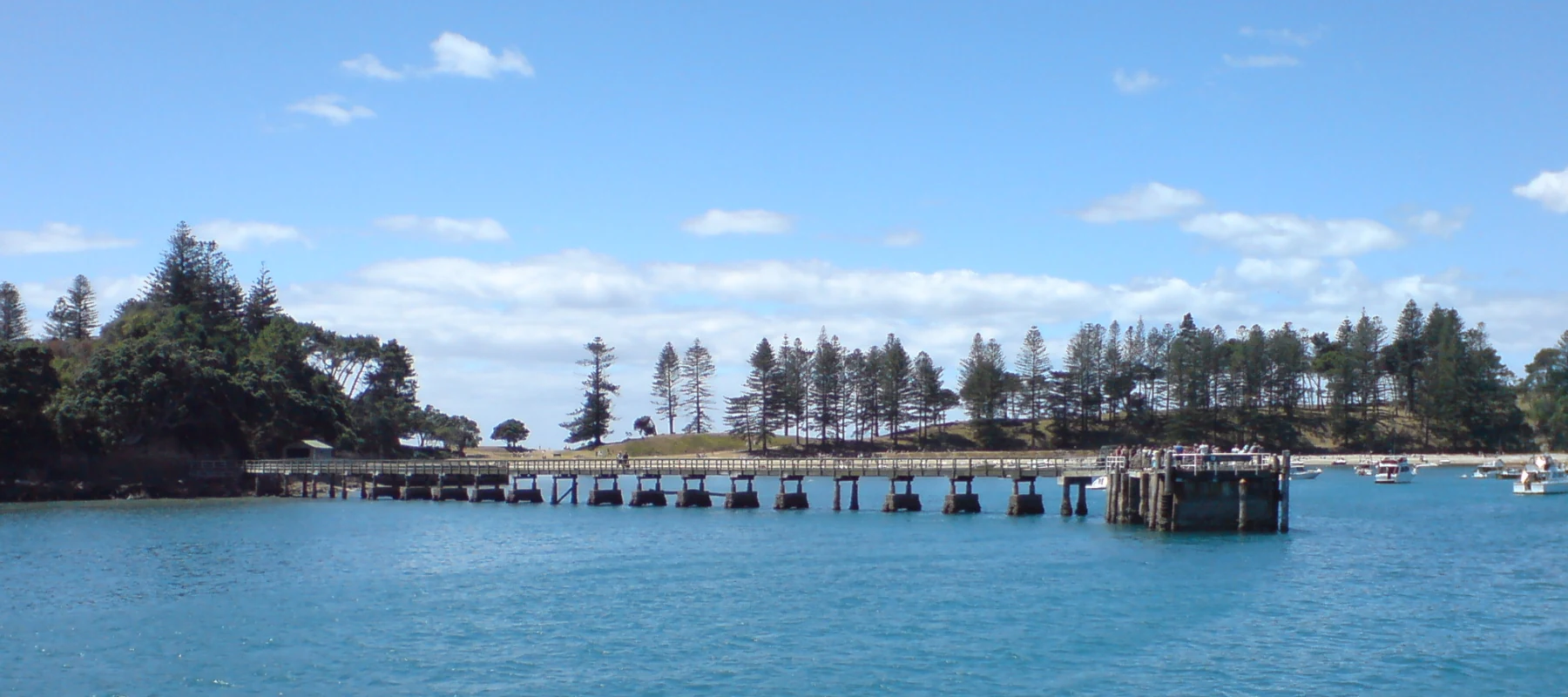 A wide, panoramic view of the wooden wharf stretching out into the calm blue water towards the island shore, with green trees and a clear blue sky in the background.