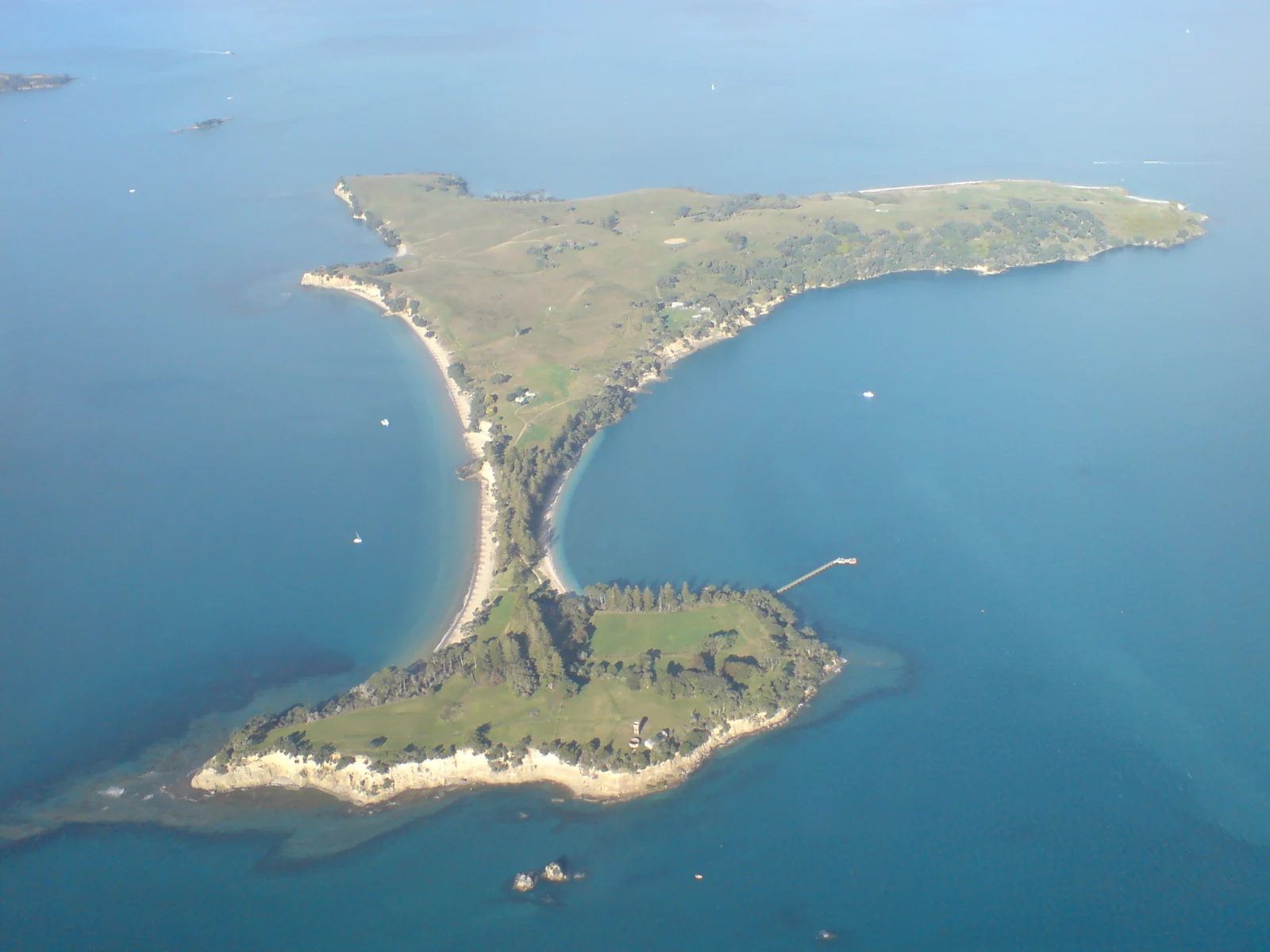 A high angle aerial view of Motuihe Island, which is a thin, green island surrounded by the blue waters of the Hauraki Gulf.