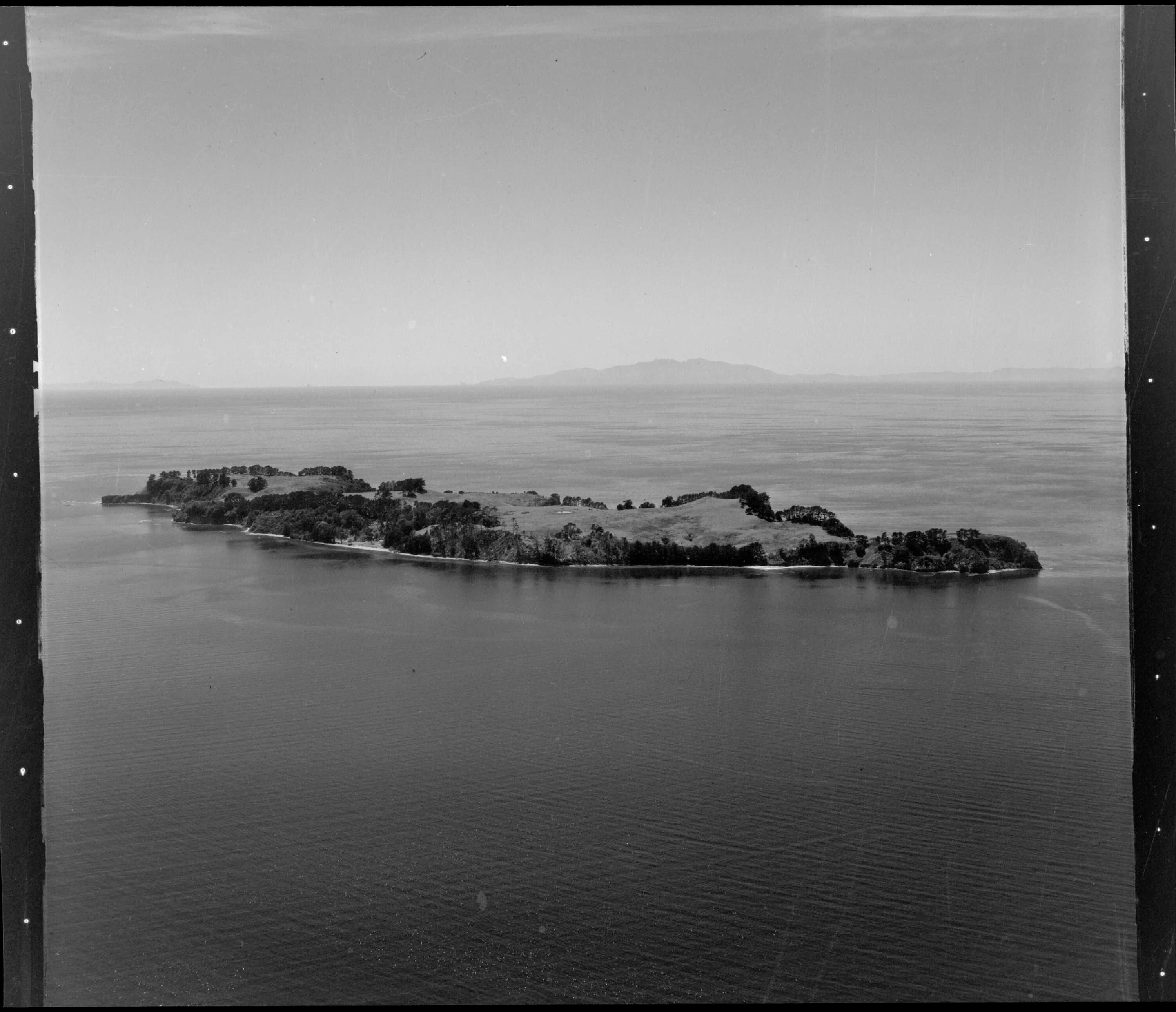 Una fotografía aérea en blanco y negro que muestra la totalidad de la masa terrestre de la isla Motuora rodeada por aguas marinas en calma.