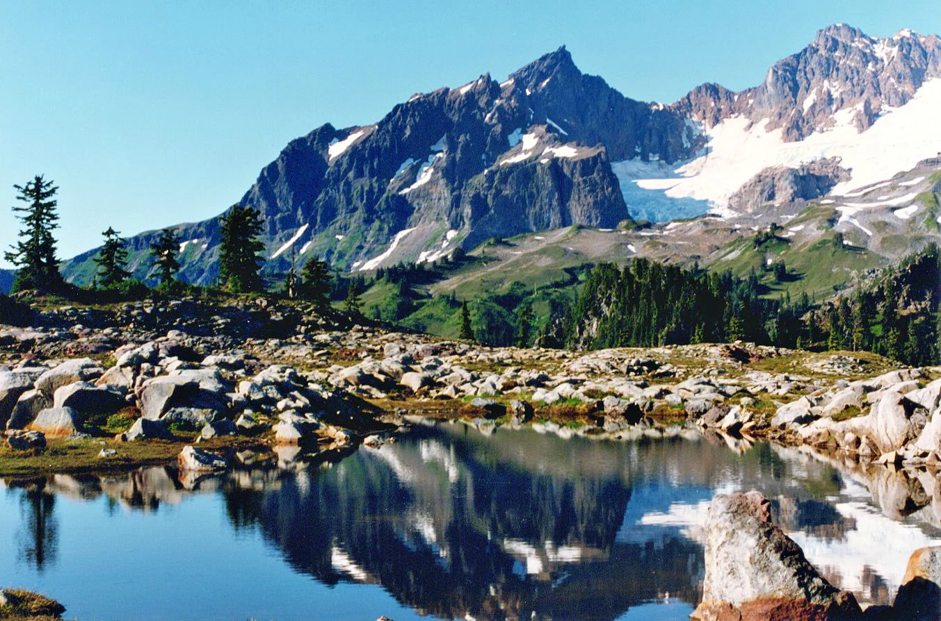 A clear mountain lake reflects the rugged, glacier-covered Black Buttes in the background, with rocky foreground terrain and small evergreen trees.