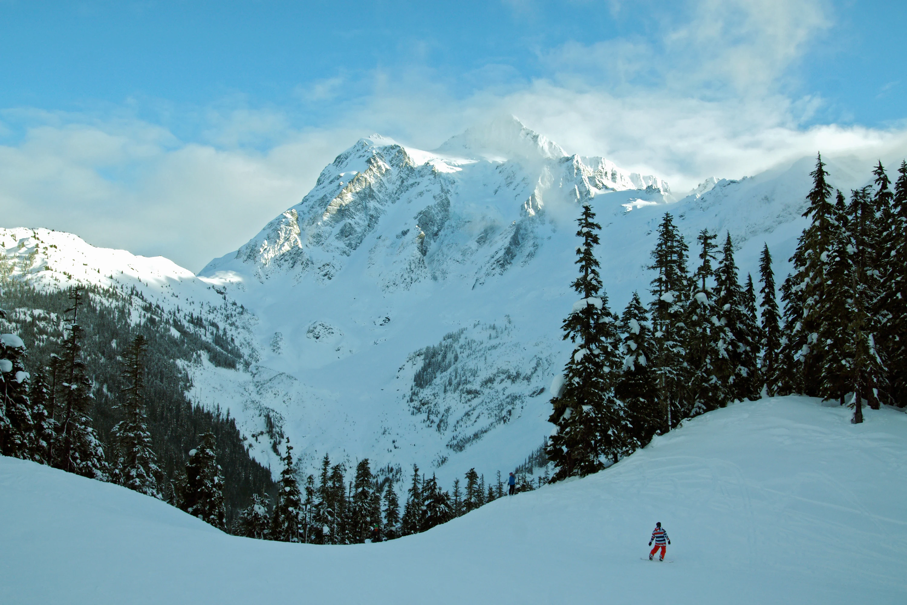 A lone snowboarder moves across a large, snow-covered landscape with a massive, jagged, snow-covered mountain peak looming in the background behind a cluster of evergreen trees.