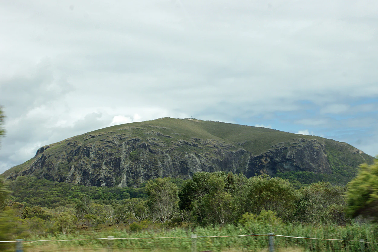 A large hill with rocky slopes and green vegetation under a cloudy sky, with trees and grass in the foreground