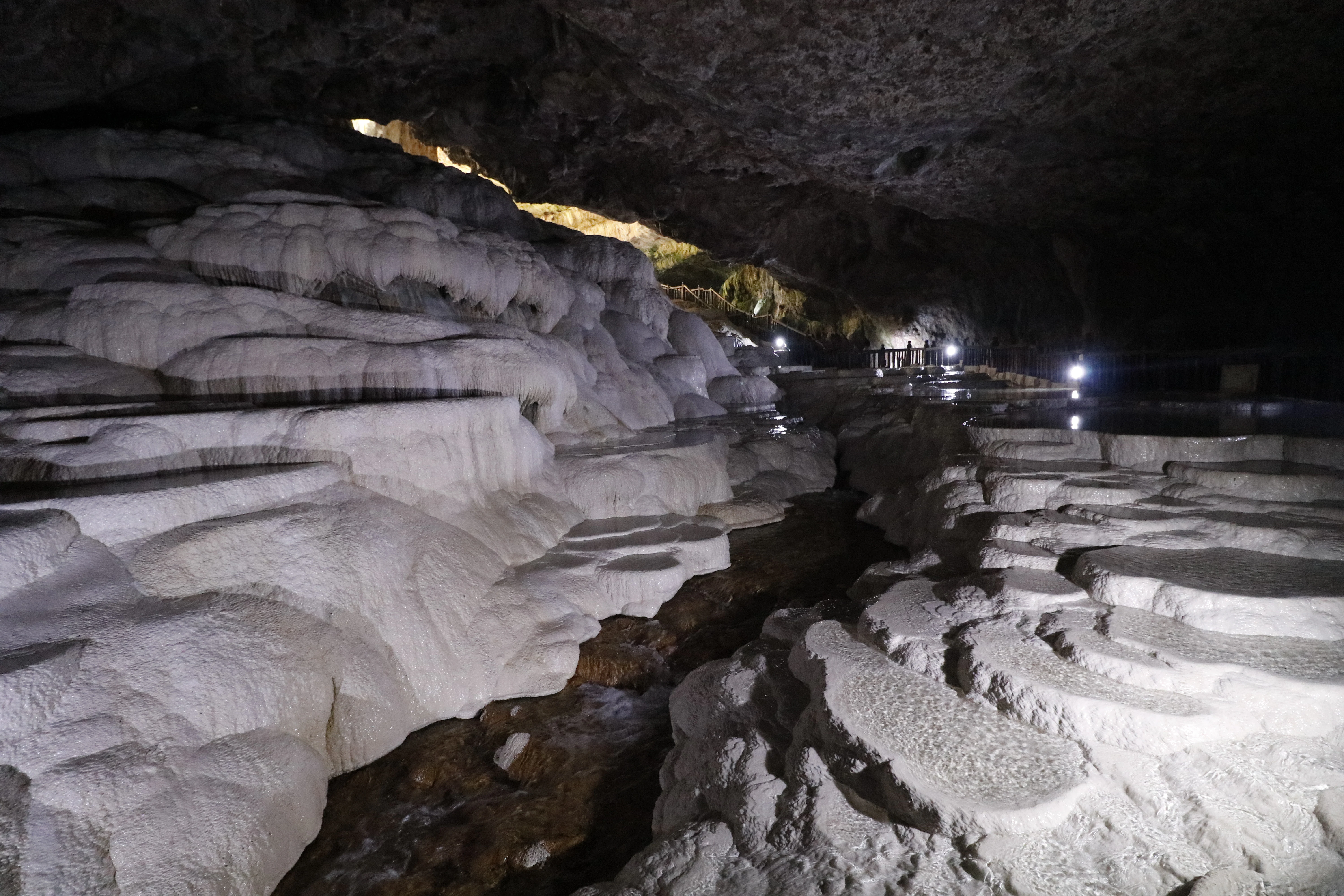 Cave interior featuring white rock formations, flowing water, and artificial lighting