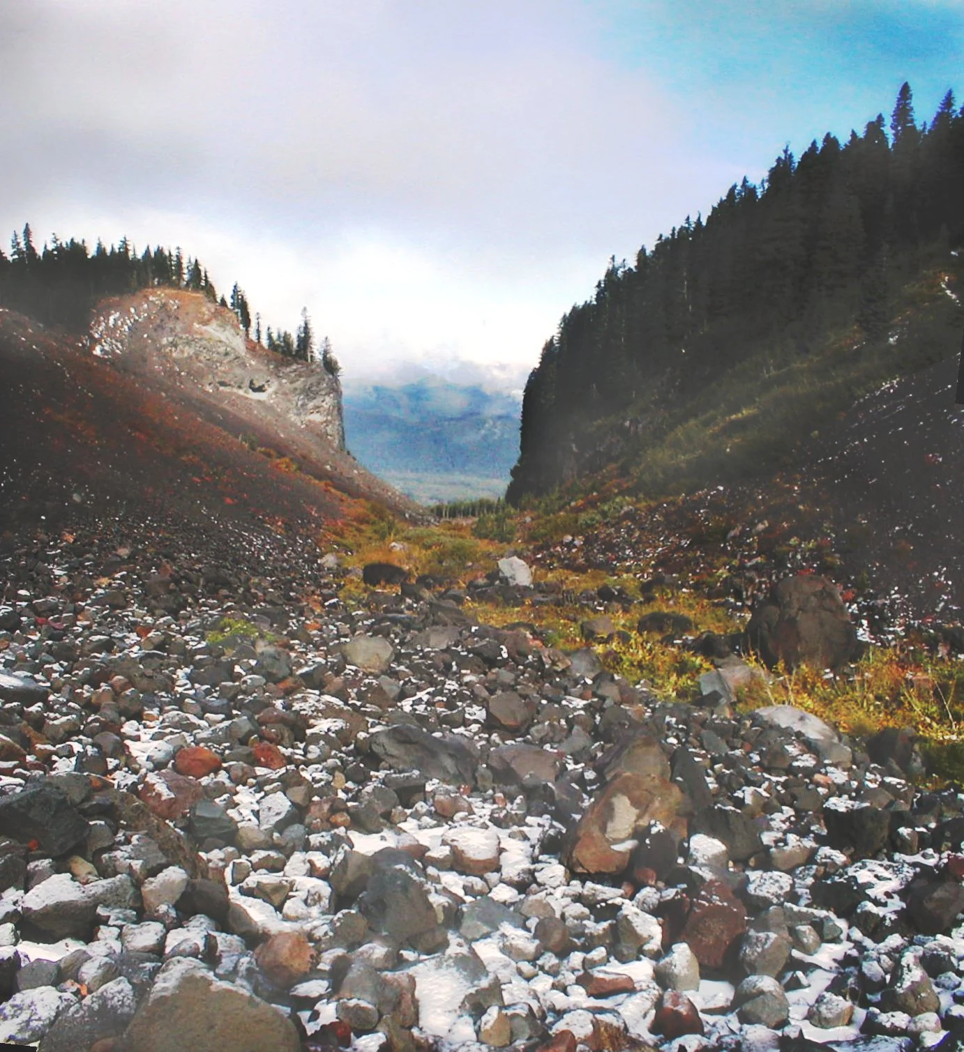 A low angle view of a rocky glacial valley with dark, steep slopes covered in volcanic rock and patches of snow, flanked by coniferous forests, leading toward a distant misty peak.