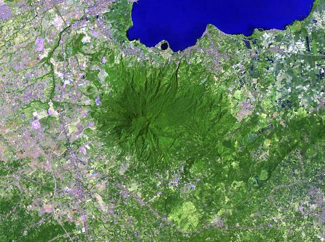 Top-down satellite imagery of Mount Makiling, appearing as a dense circular area of green forest surrounded by agricultural and urban land, with a large blue body of water to the north.