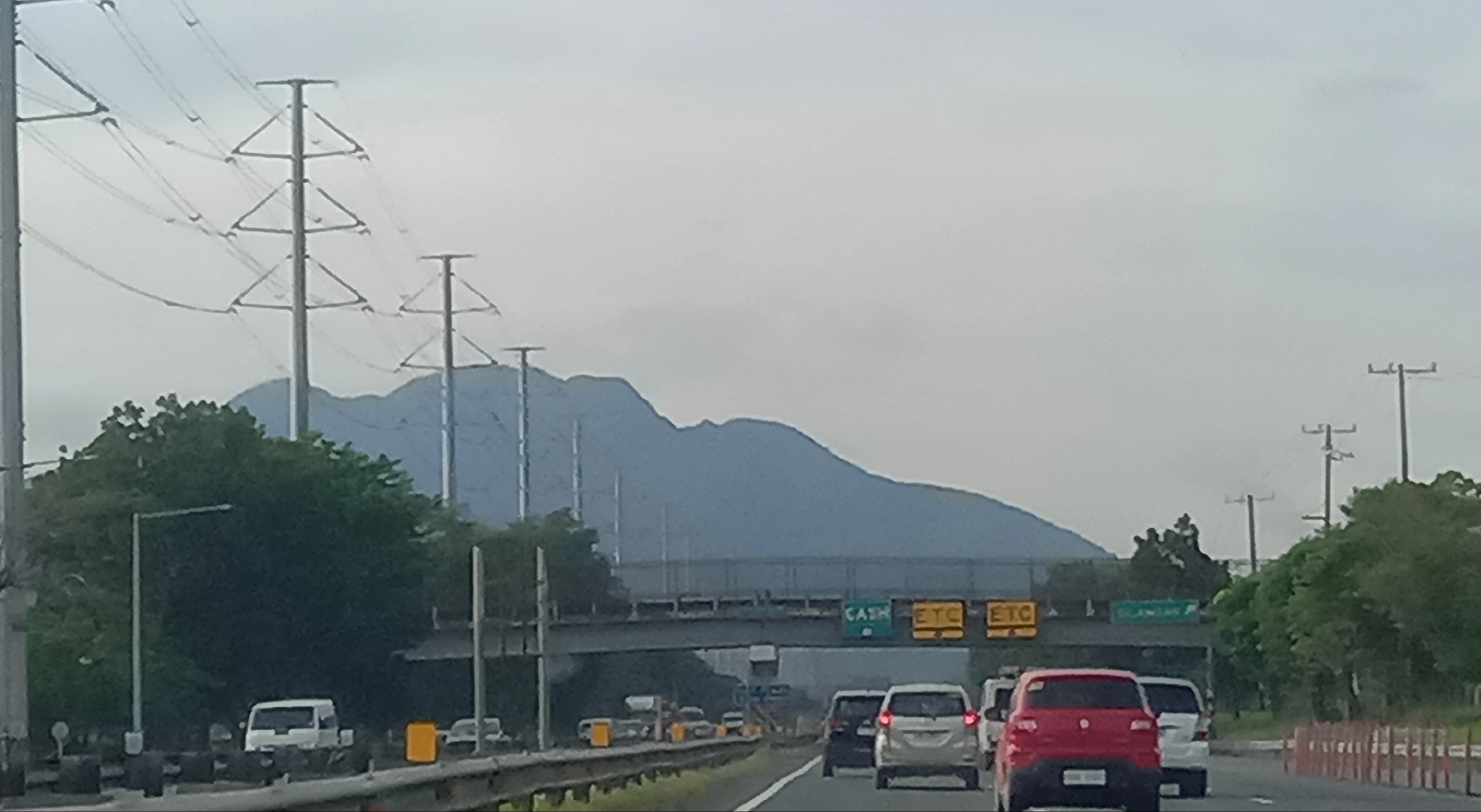 A distant view of the Mount Makiling dormant volcano silhouette rising above a highway with cars and power line towers in the foreground.