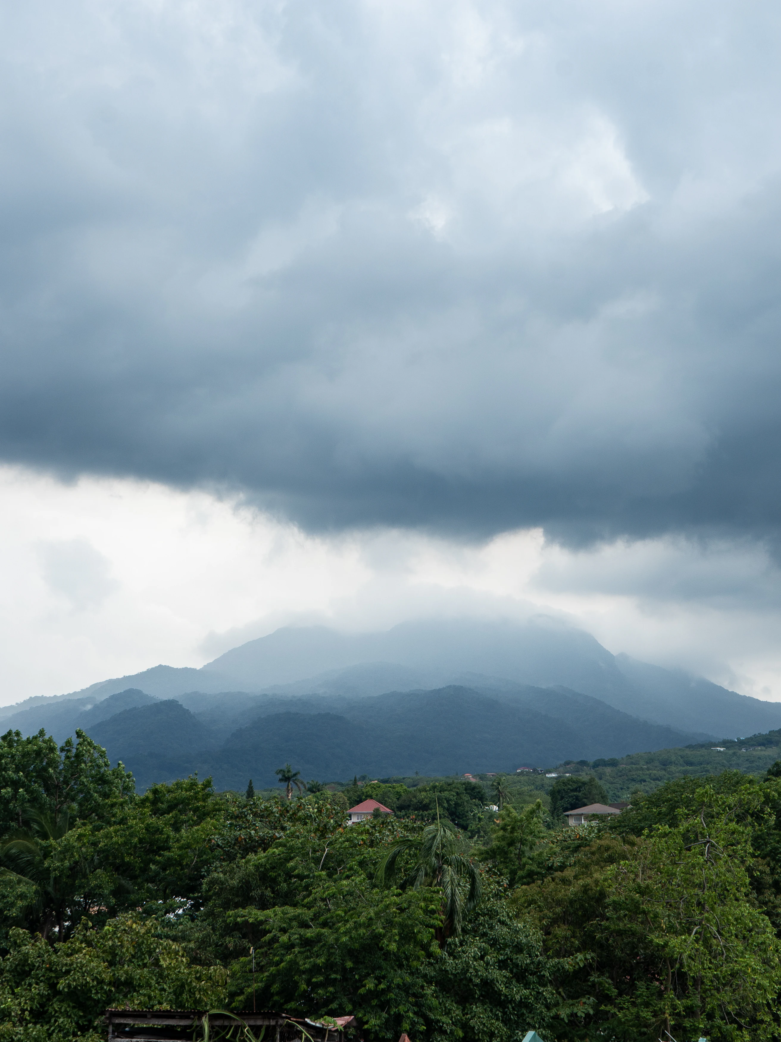 A wide view of Mount Makiling, an inactive stratovolcano covered in forest, appearing beneath heavy, dark rain clouds.
