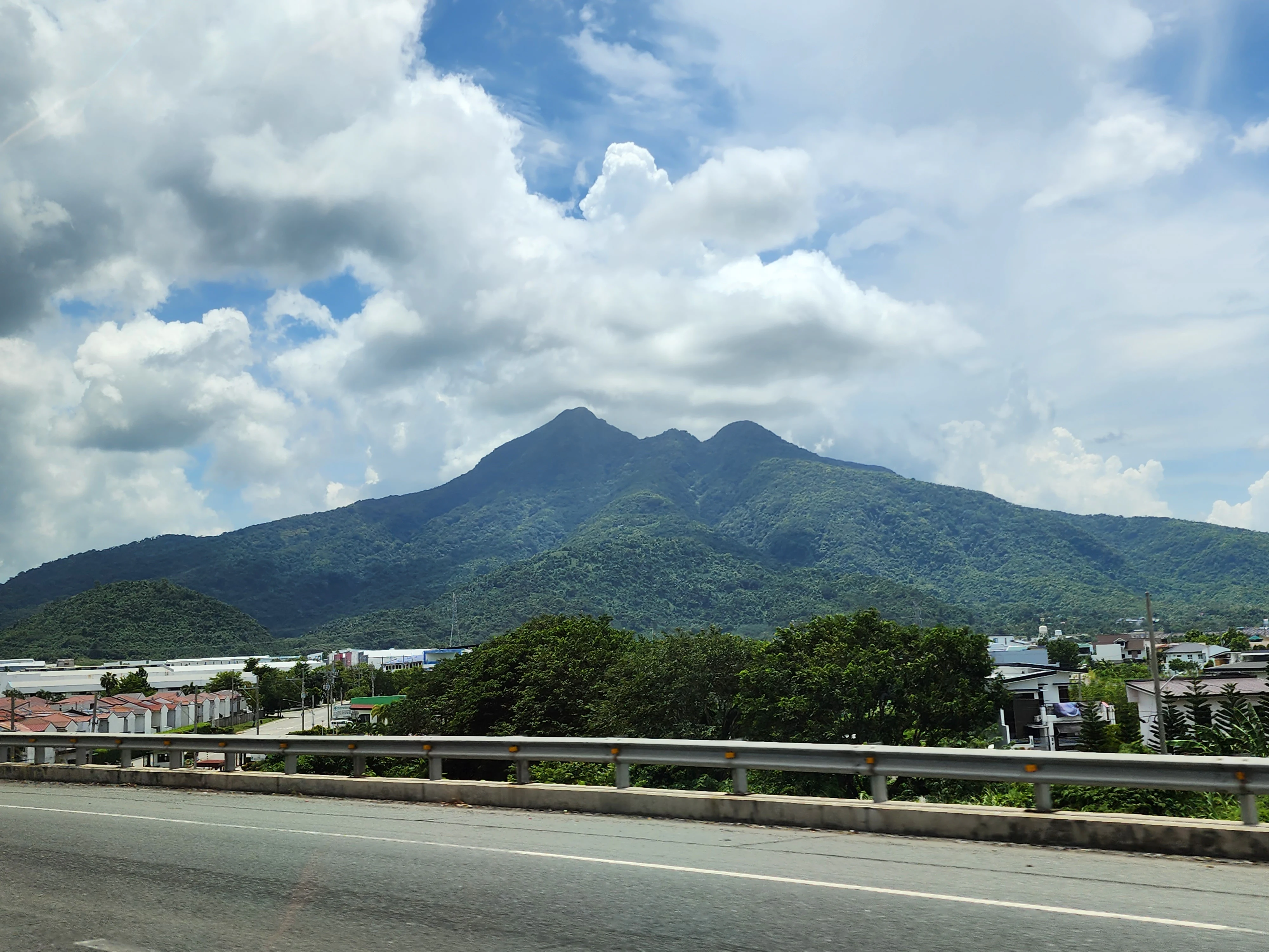A wide view of the inactive stratovolcano Mount Makiling, covered in dense green forest, under a partly cloudy sky, with a highway guardrail and road in the foreground.