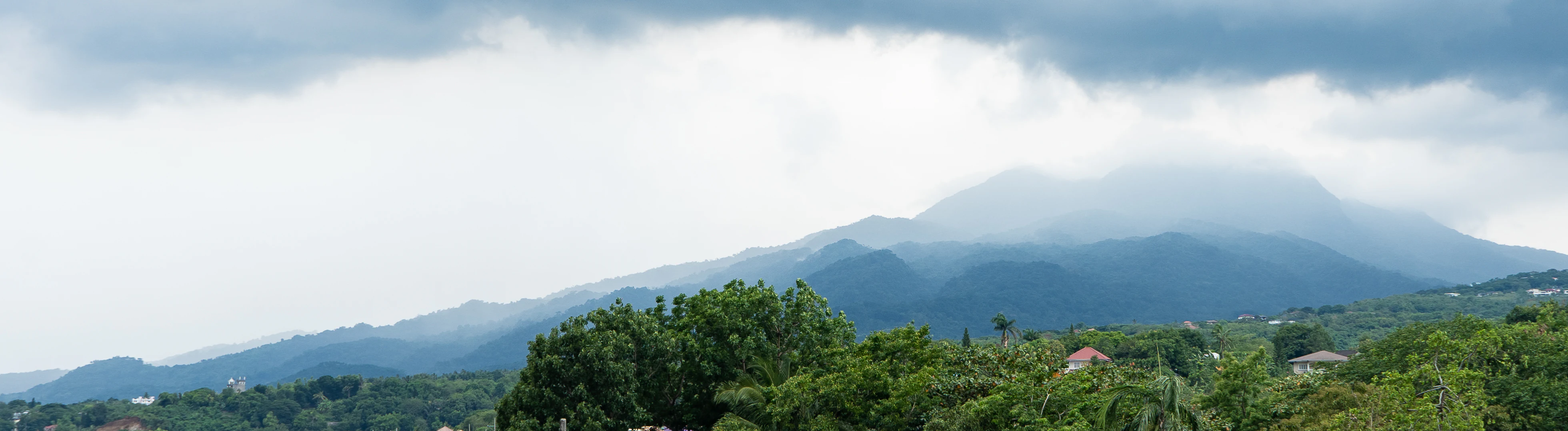 A wide shot of the Mount Makiling stratovolcano, partly obscured by clouds and mist, rising above a foreground of lush green trees and distant residential buildings.
