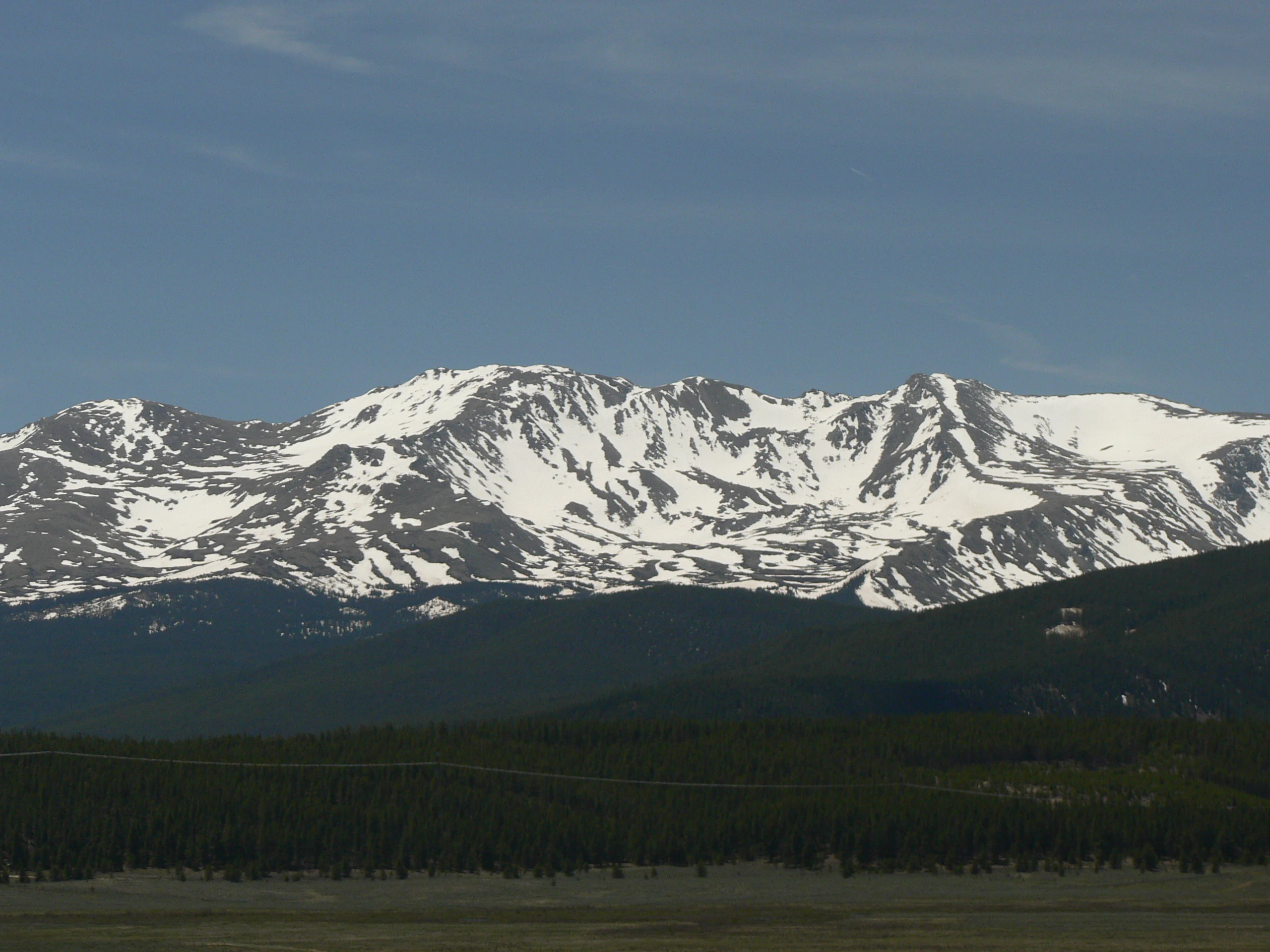 A wide landscape view of a prominent mountain range with snowy peaks, rising above a dark forested slope and a grassy foreground under a blue sky.