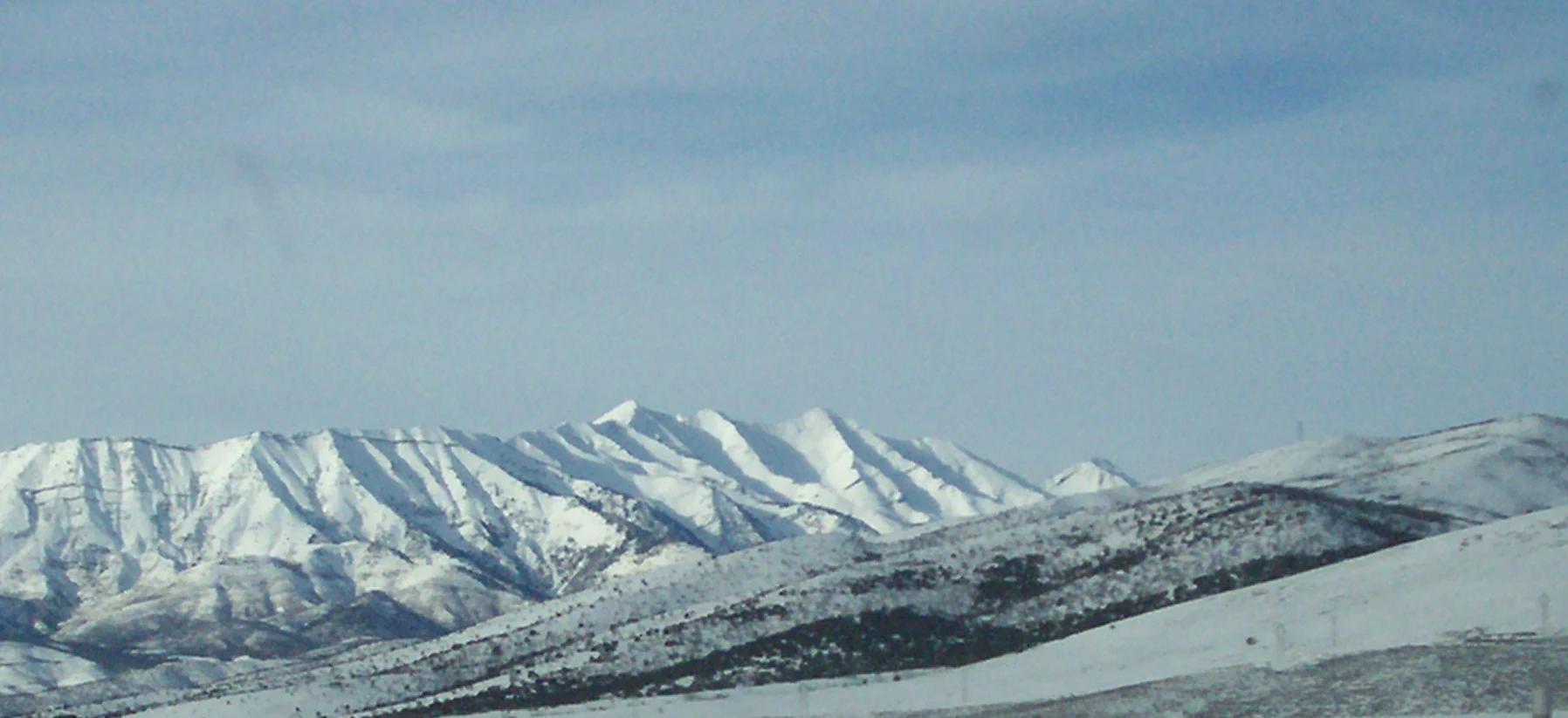 A wide, horizontal view of snow-covered mountain peaks under a clear, pale blue sky.