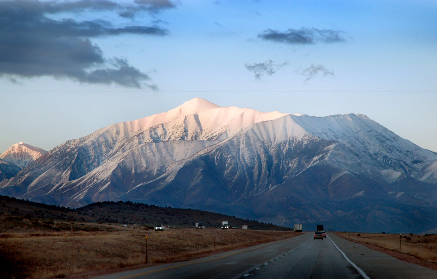 A wide, eye level shot of a large, snow dusted mountain peak rising above a desert landscape with a highway in the foreground.