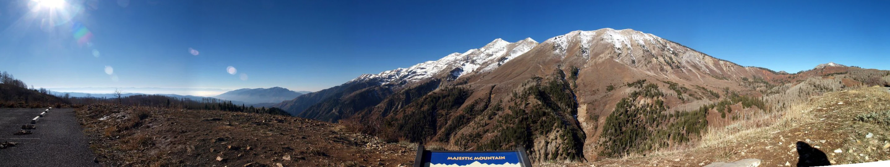 A wide, panoramic view of a large, snow-dusted mountain range under a clear blue sky, seen from a high vantage point with a small interpretive sign in the foreground.