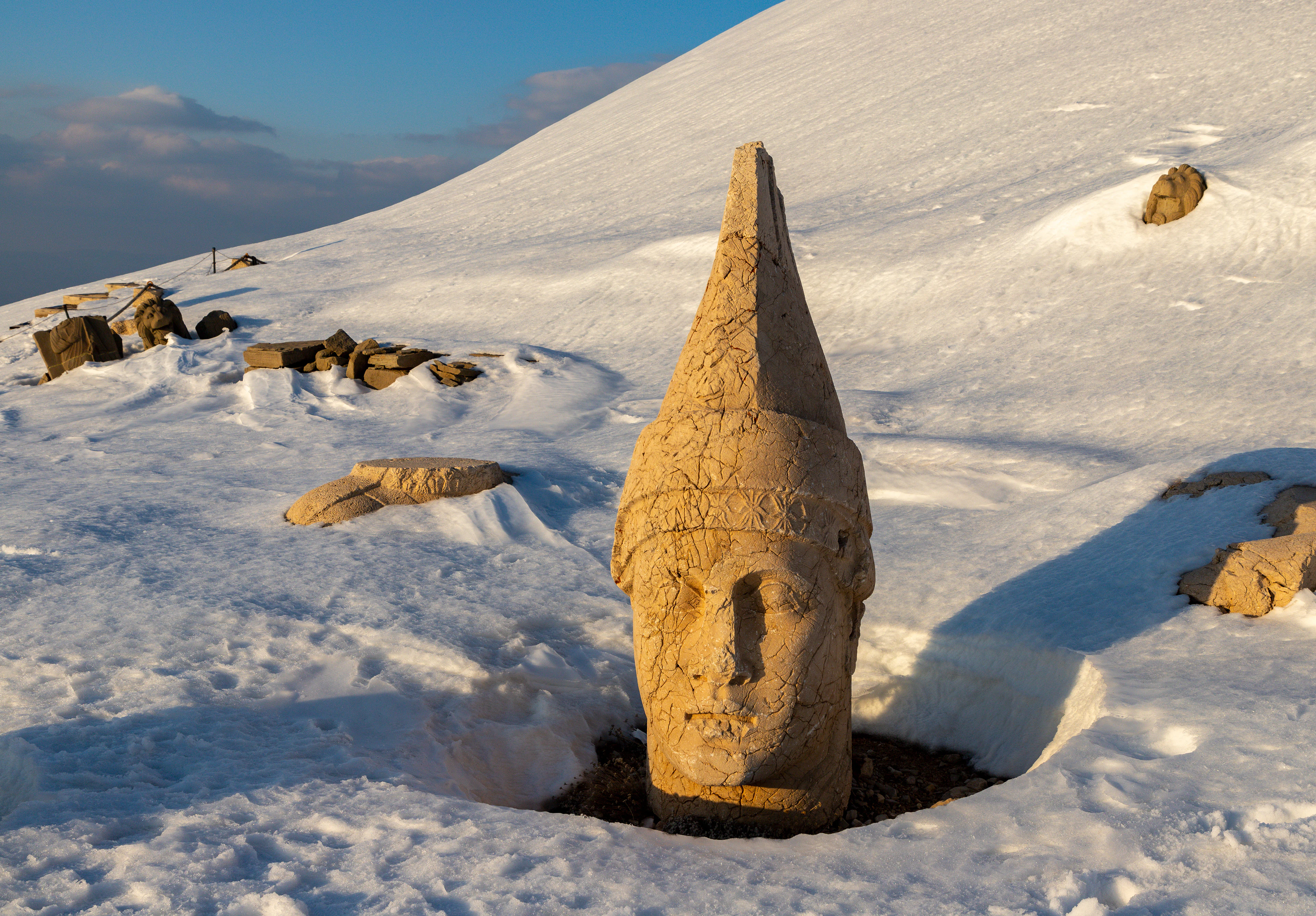 Large stone head statue partially buried in snow on a mountain slope with rocky outcrops and clear blue sky.