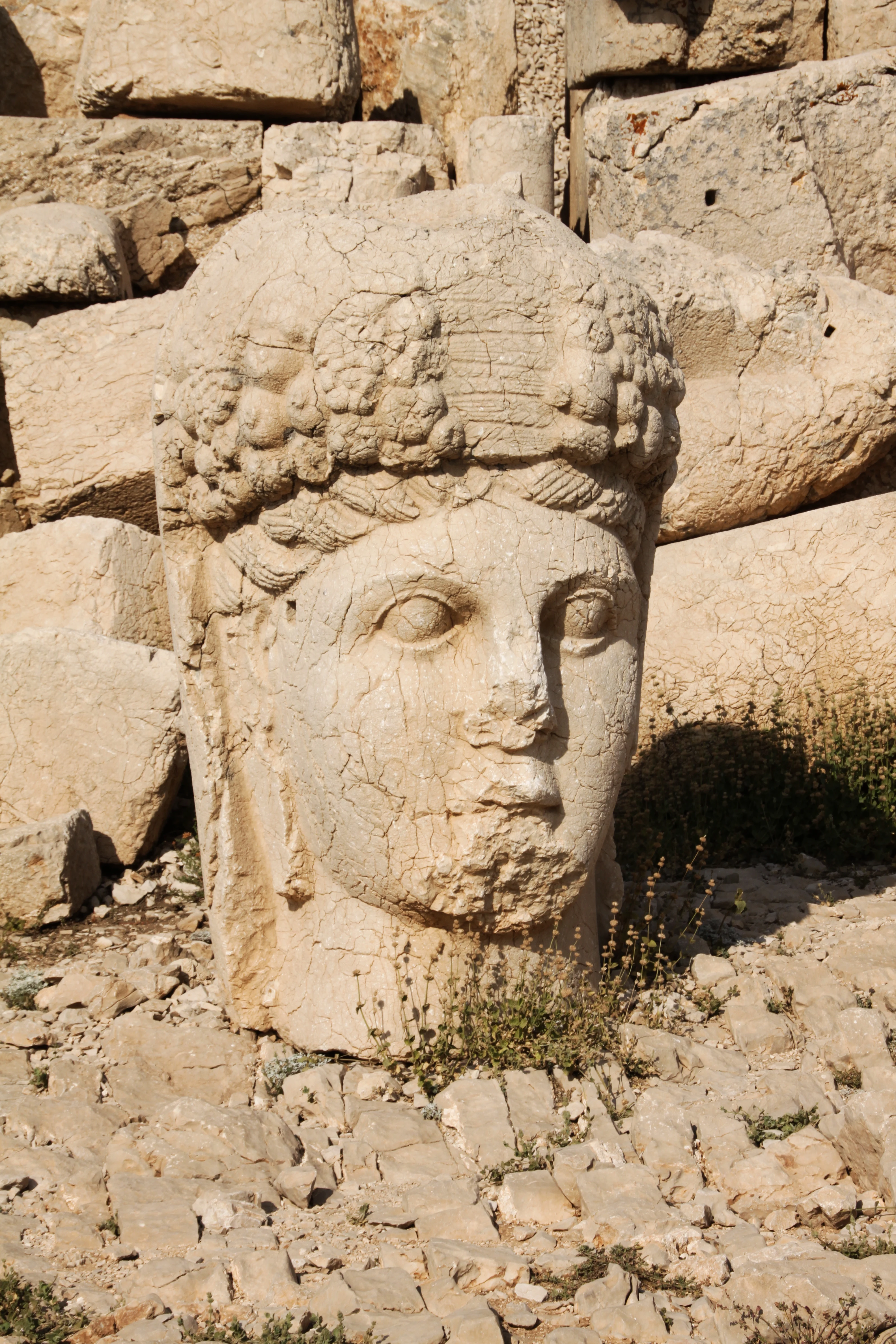 Ancient stone head sculpture with cracked surface surrounded by large rocks and sparse vegetation
