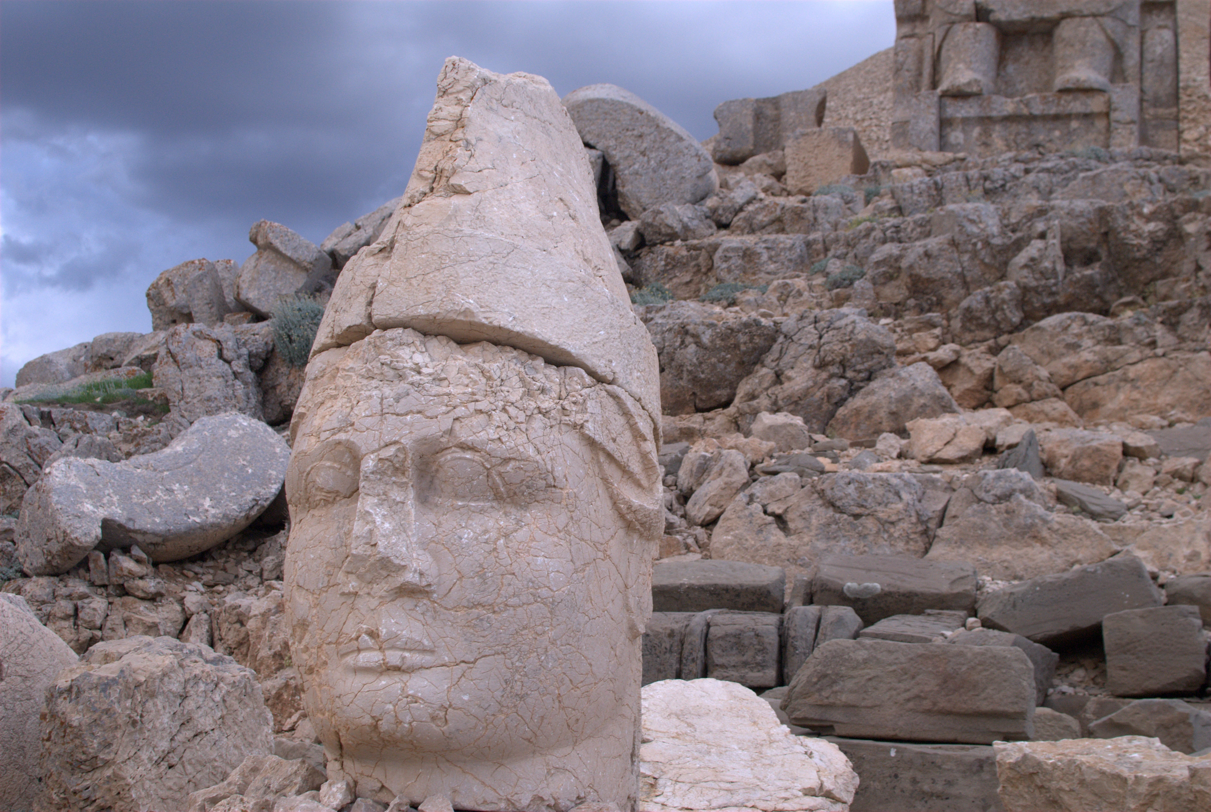 Large ancient stone head sculpture with a conical hat, surrounded by rocky terrain and ruins, under a cloudy sky