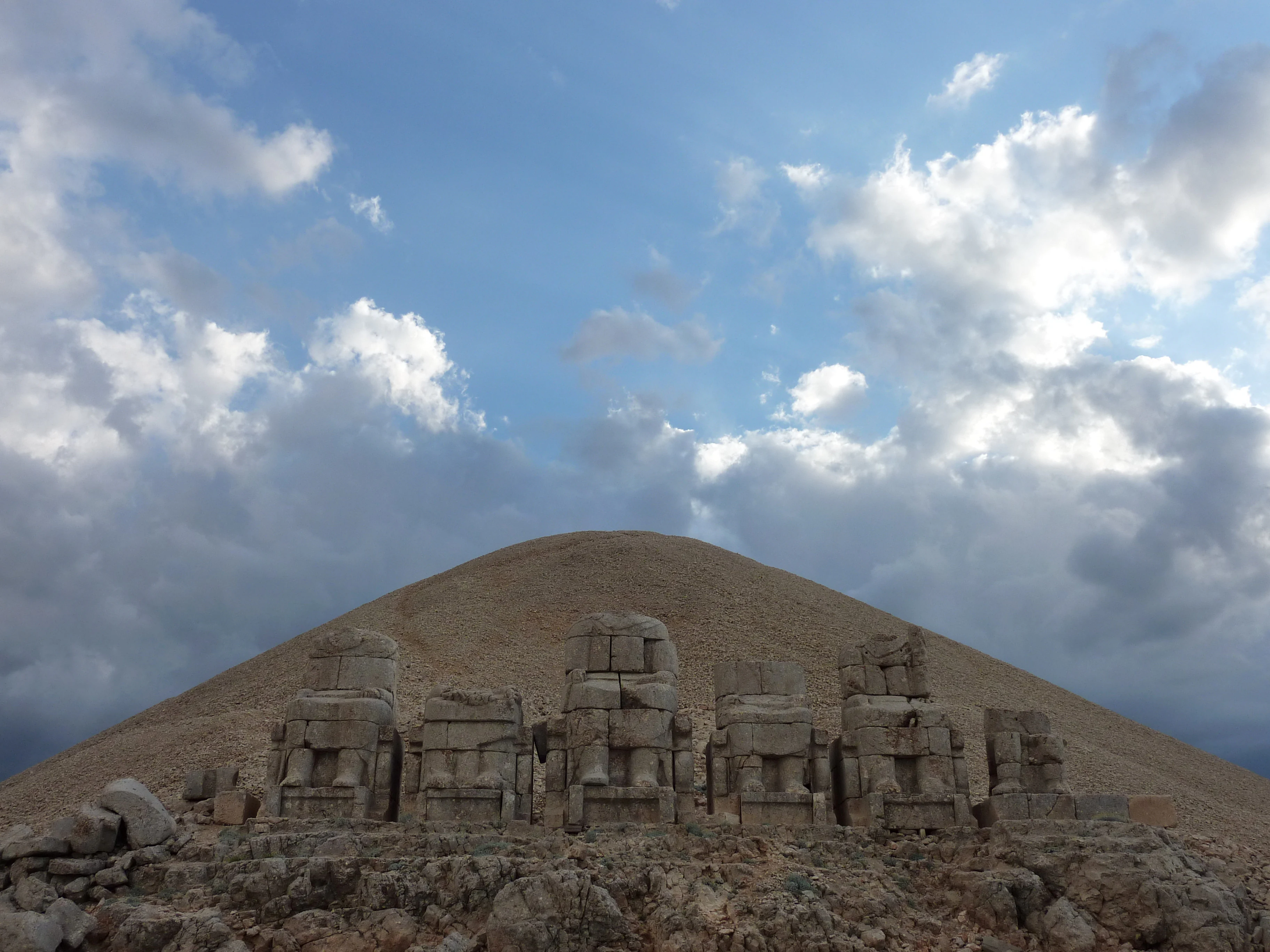 Stone statues at the summit of Mount Nemrut with a mountain backdrop under a cloudy sky