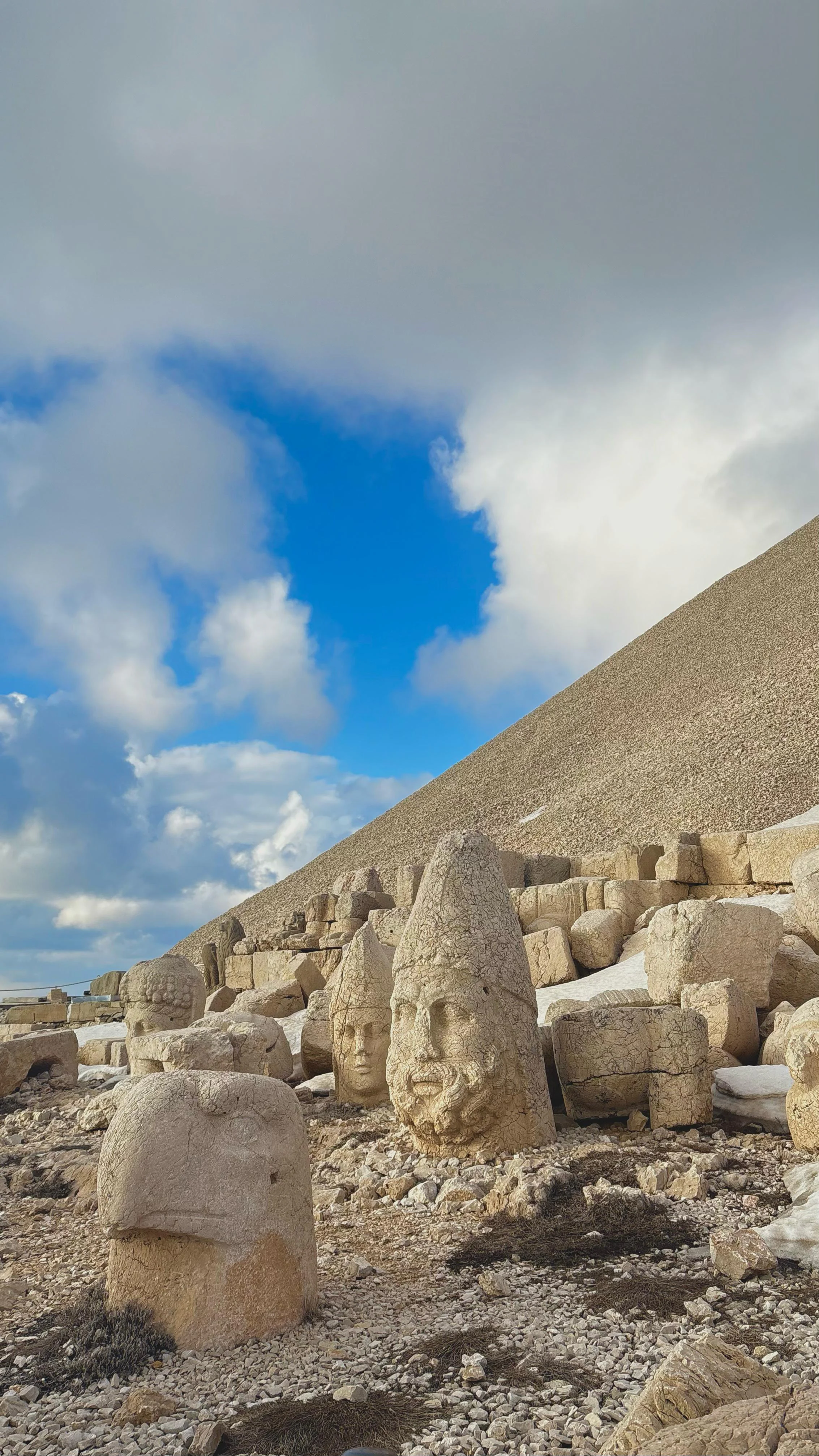Stone statues and fragments on rocky terrain with a mountain slope in the background under a partly cloudy sky.