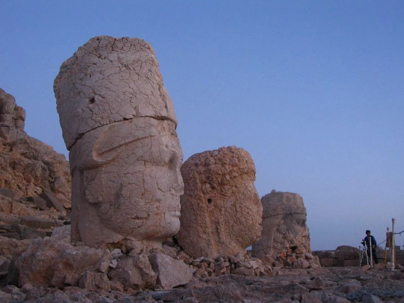 Large ancient stone heads on rocky terrain under clear sky at dusk with a person standing nearby