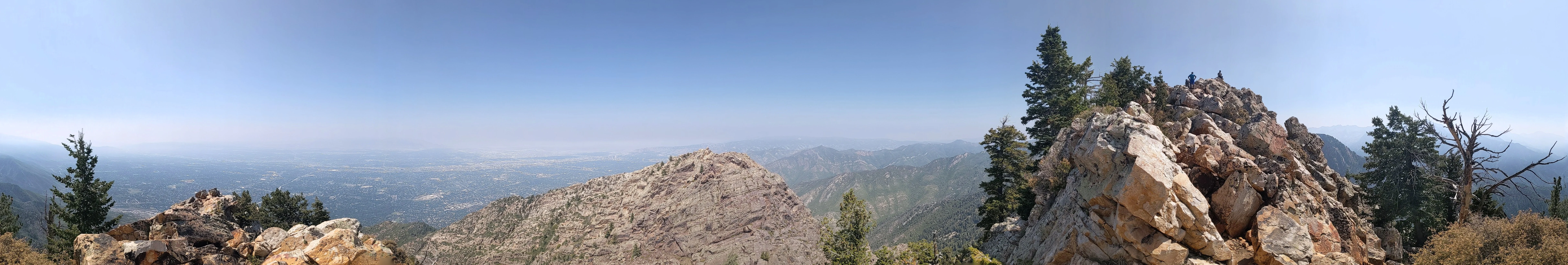Panoramic view from a rocky mountain peak overlooking layers of lower mountain ridges and a hazy valley under a clear blue sky.