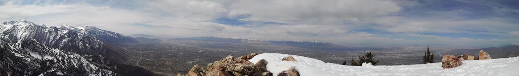 Panoramic view from a snowy mountain peak looking out over a wide valley under a cloudy sky.