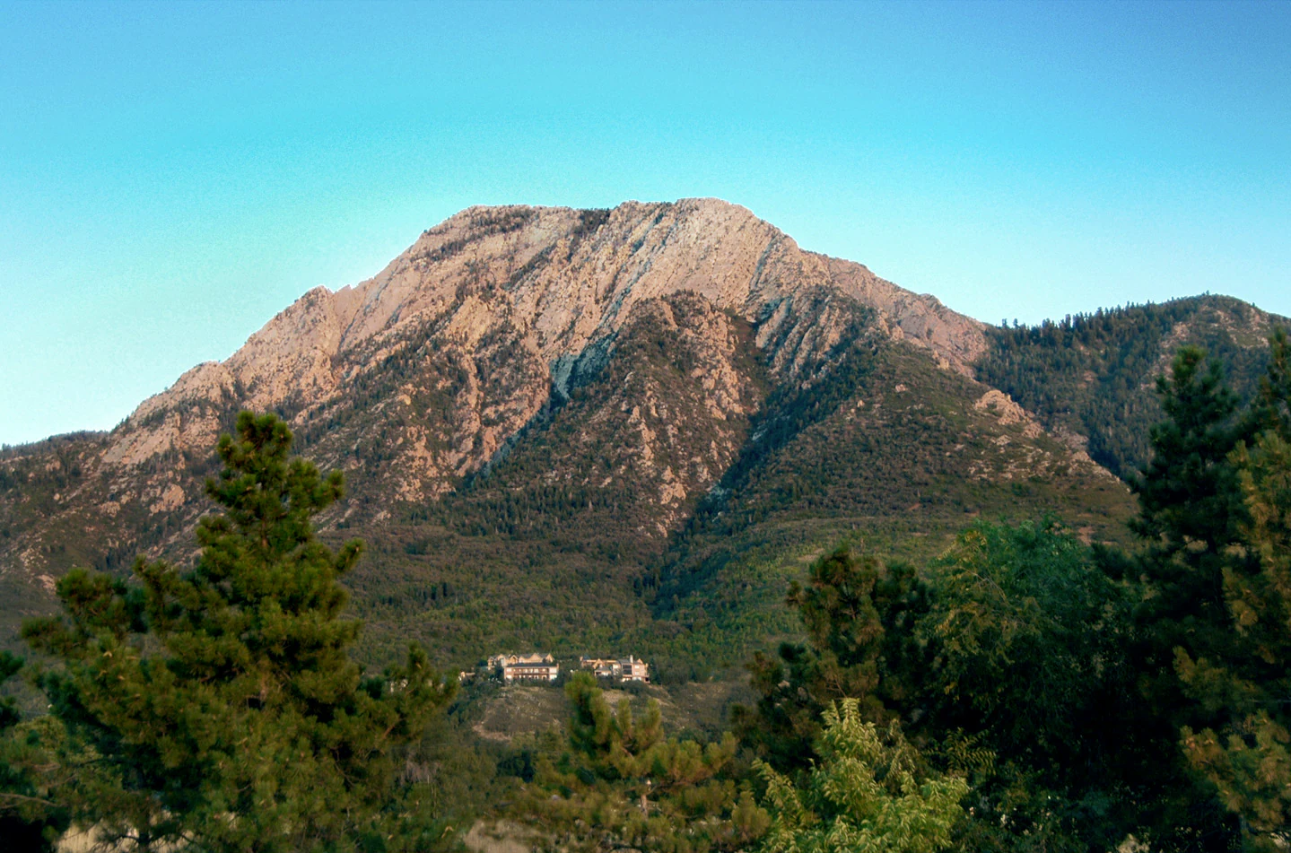 A large, rugged, tan and grey mountain peak rises above a forested slope under a clear blue sky.
