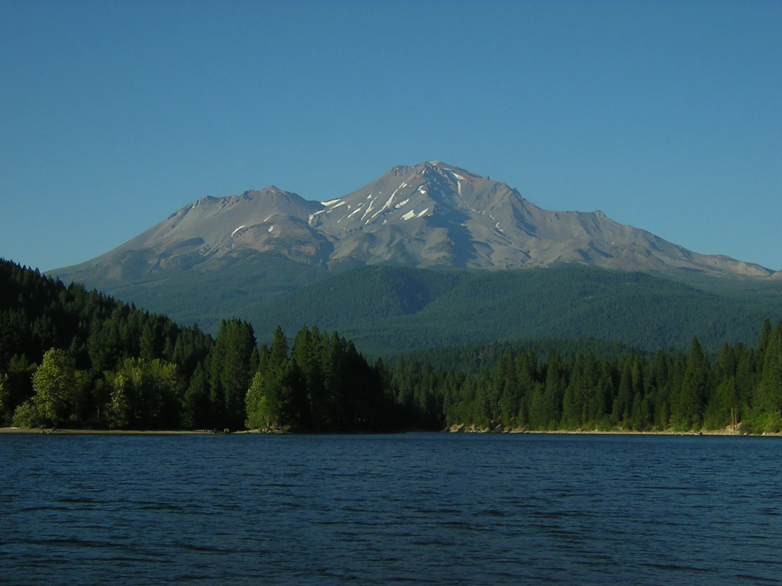 A wide view of the large, snow-dusted peak of Mount Shasta rising above a dense pine forest and the dark blue water of a lake.