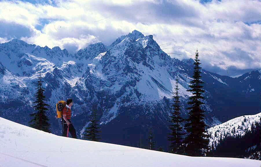 A person in winter gear with a backpack hiking across a snow-covered mountainside; tall, snow-dusted coniferous trees are visible, and jagged, snow-covered mountains rise in the distance under a partly cloudy sky.