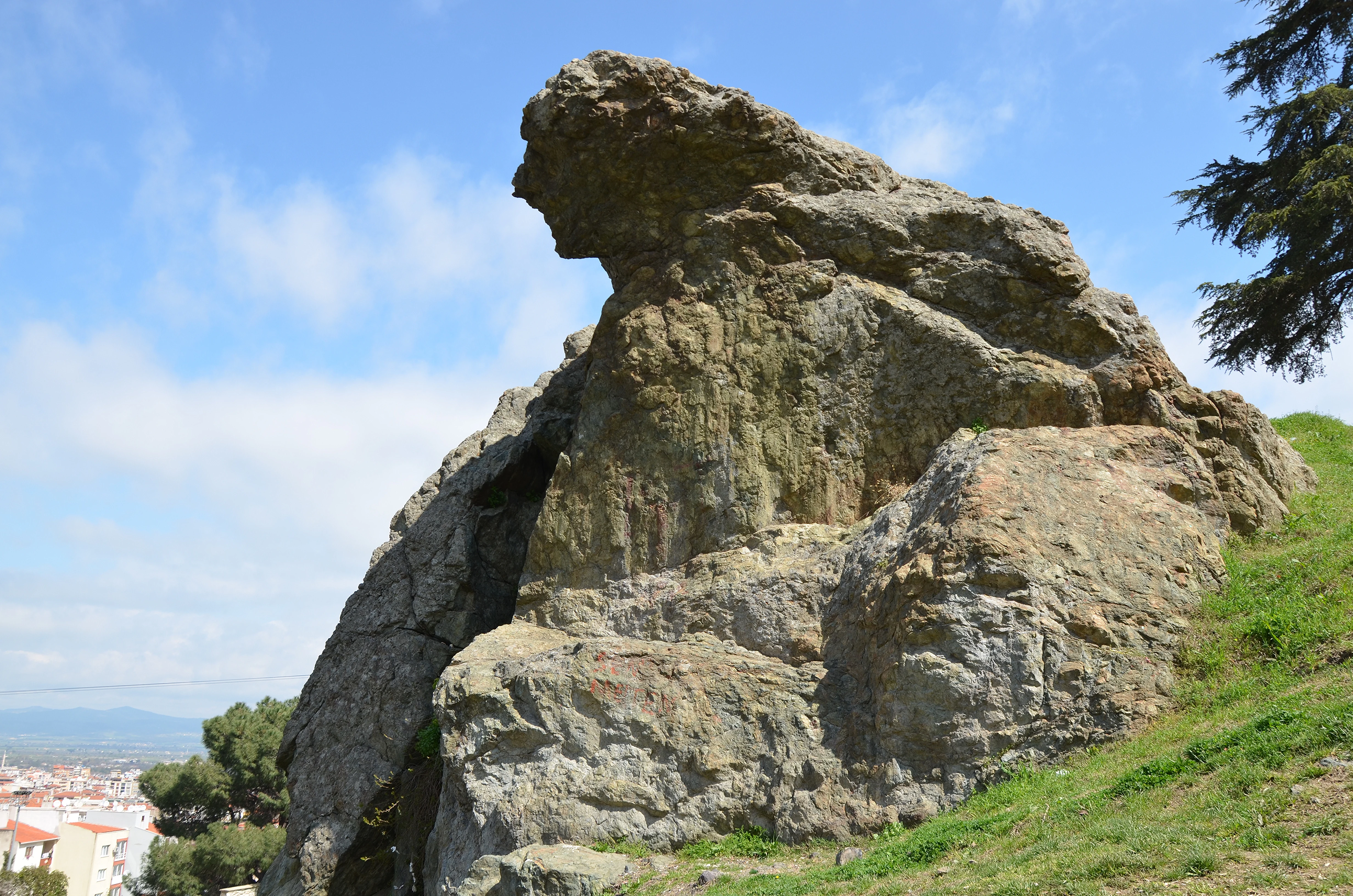 Large rock formation on a grassy hillside under a clear blue sky, with buildings visible in the distance