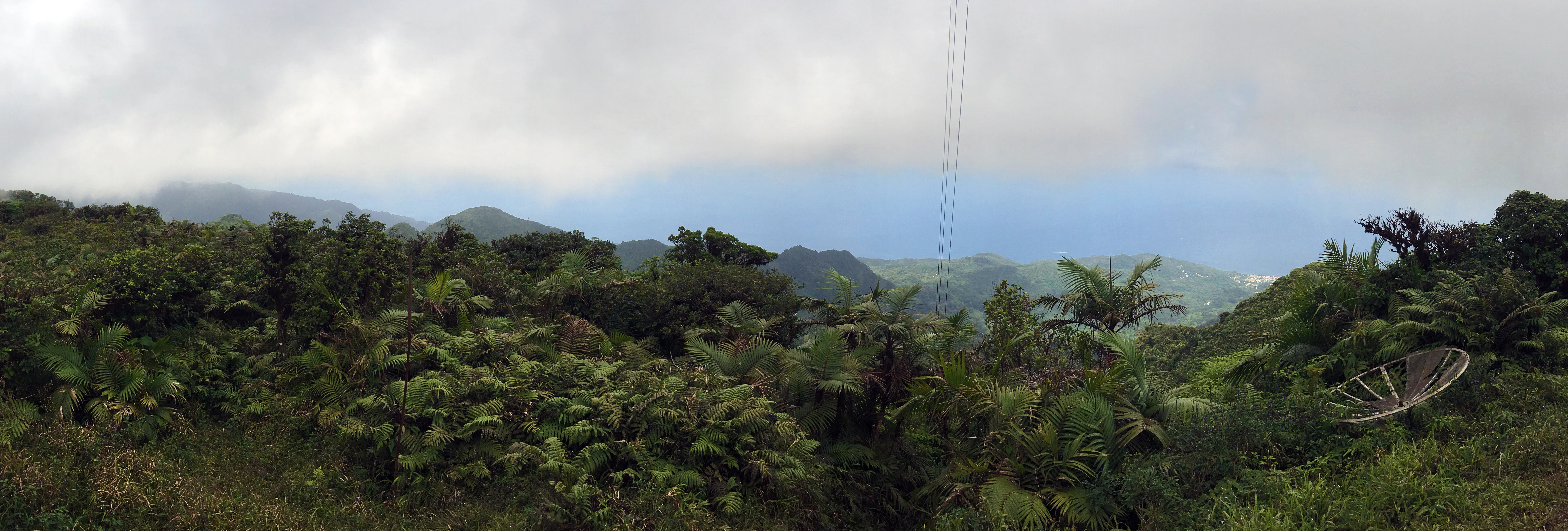 A wide, panoramic view of a lush, green mountain summit covered in dense tropical forest, with a satellite dish visible on the right and clouds clinging to the distant peaks.