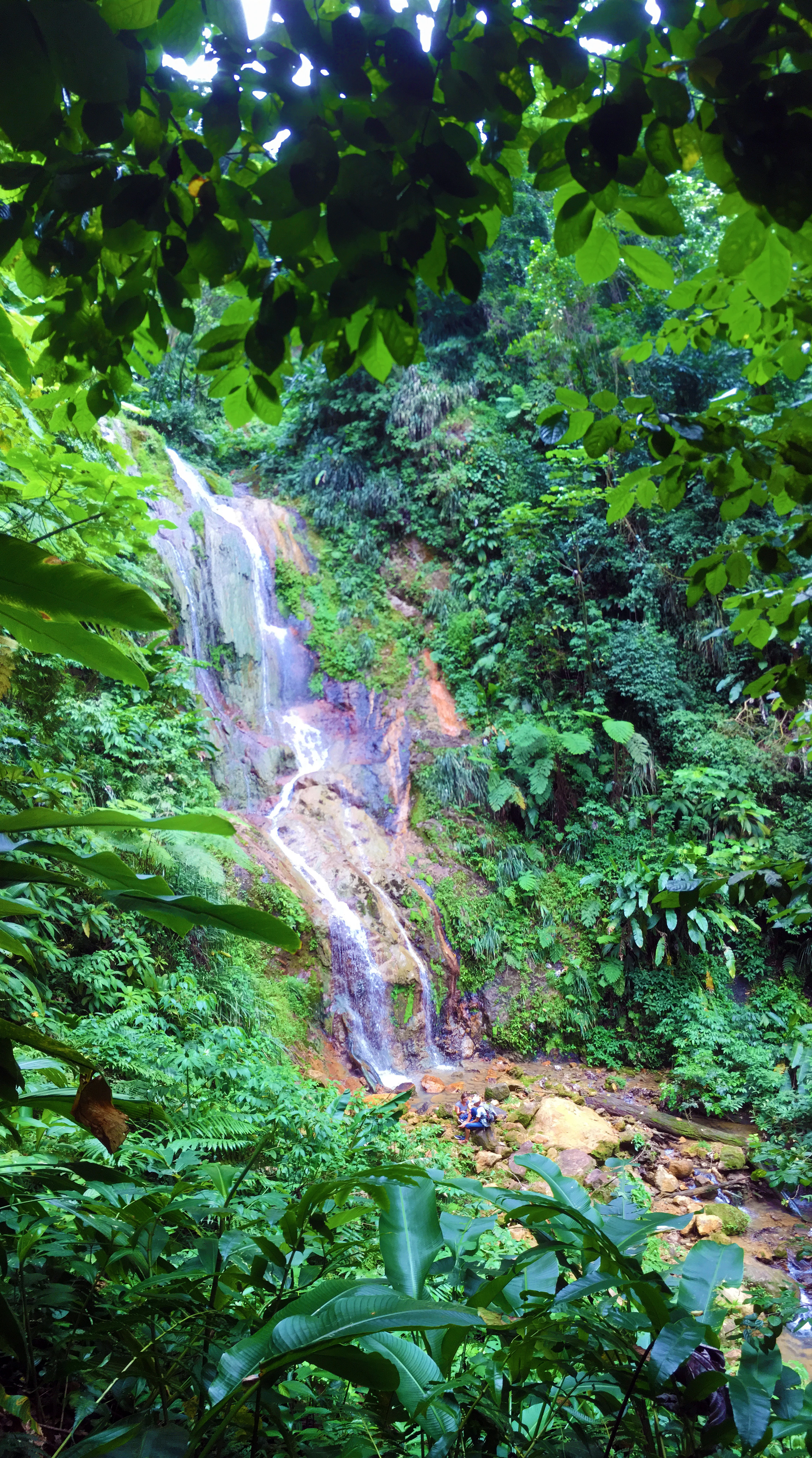 A multi-tiered waterfall cascading down a rocky, mossy slope surrounded by dense, vibrant green tropical vegetation.
