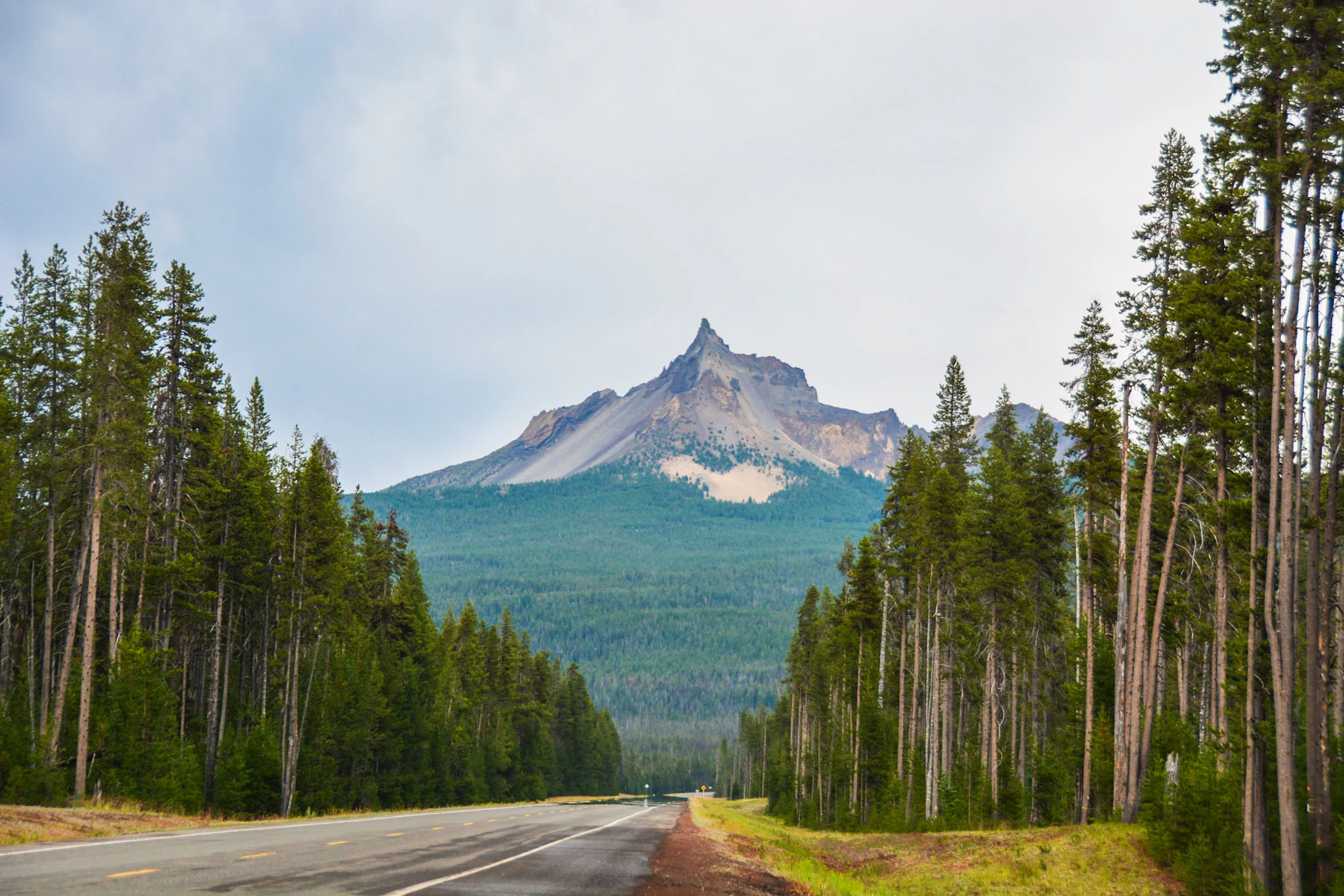 A paved road leads toward the pointed peak of Mount Thielsen, surrounded by dense coniferous forest under a cloudy sky.
