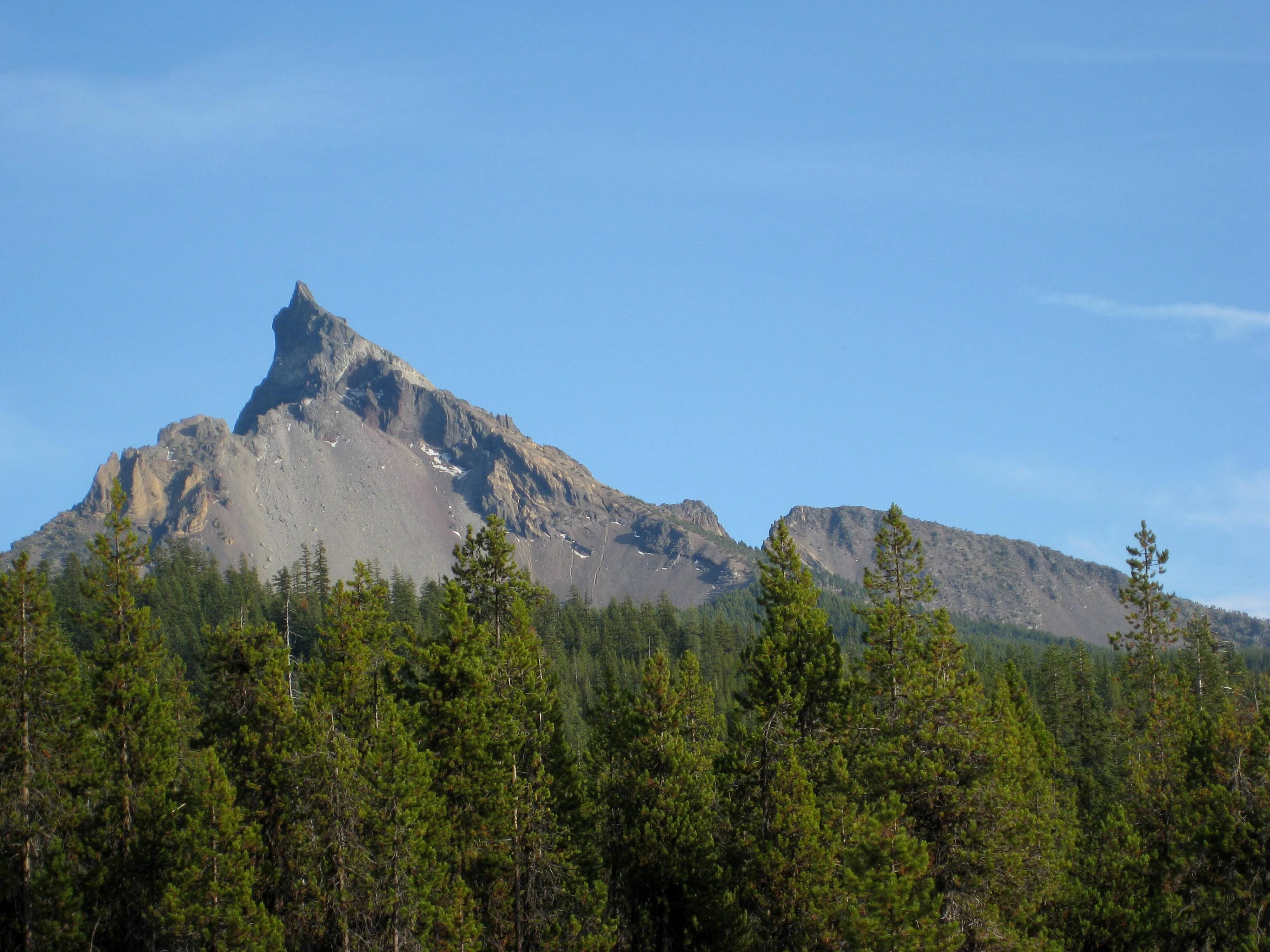 A clear view of the craggy, pointed summit of Mount Thielsen rising above a dense coniferous forest under a clear blue sky.