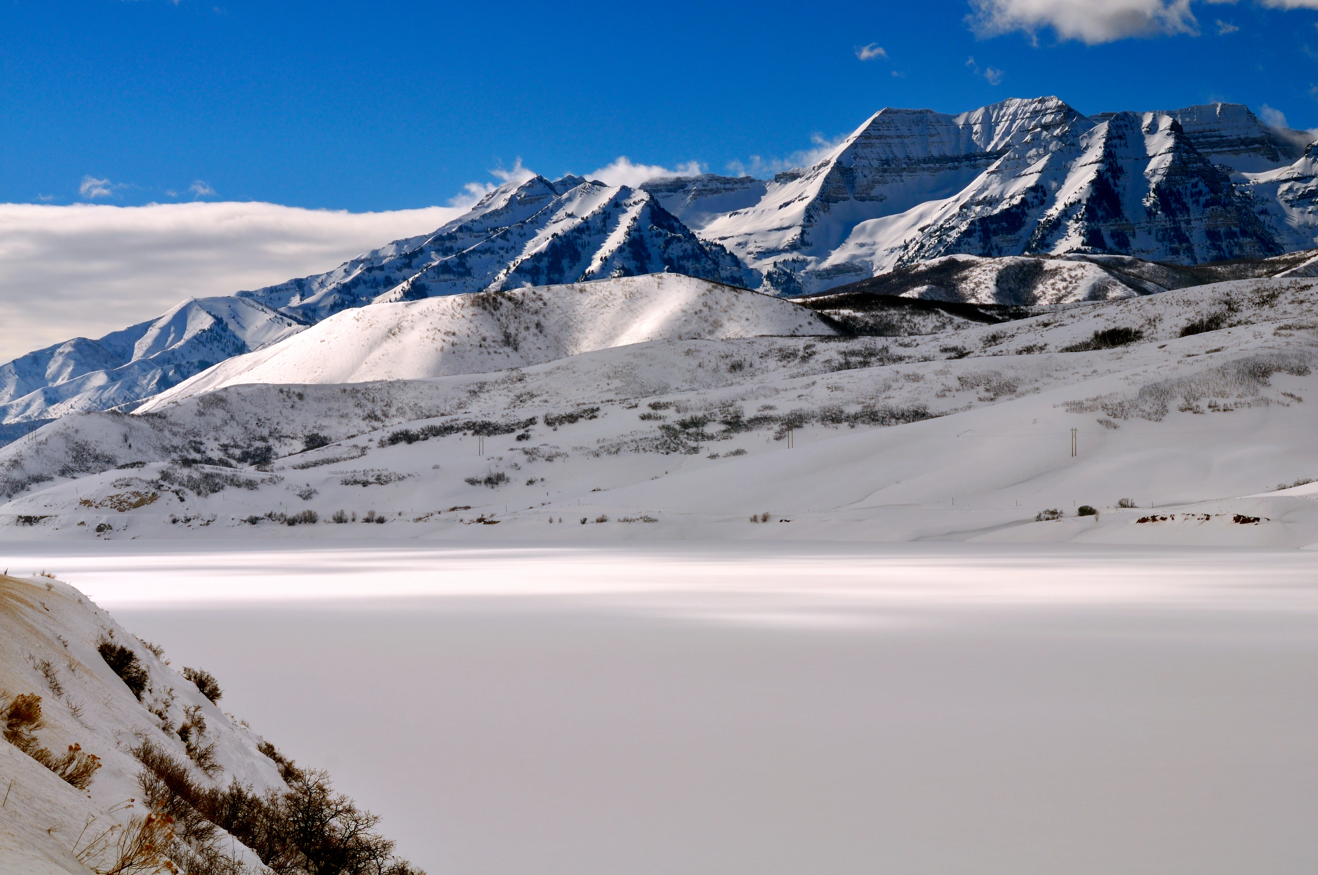 A wide, snowy landscape featuring the rugged, snow-capped peaks of Mount Timpanogos under a clear blue sky, with a large snow-covered foreground area.