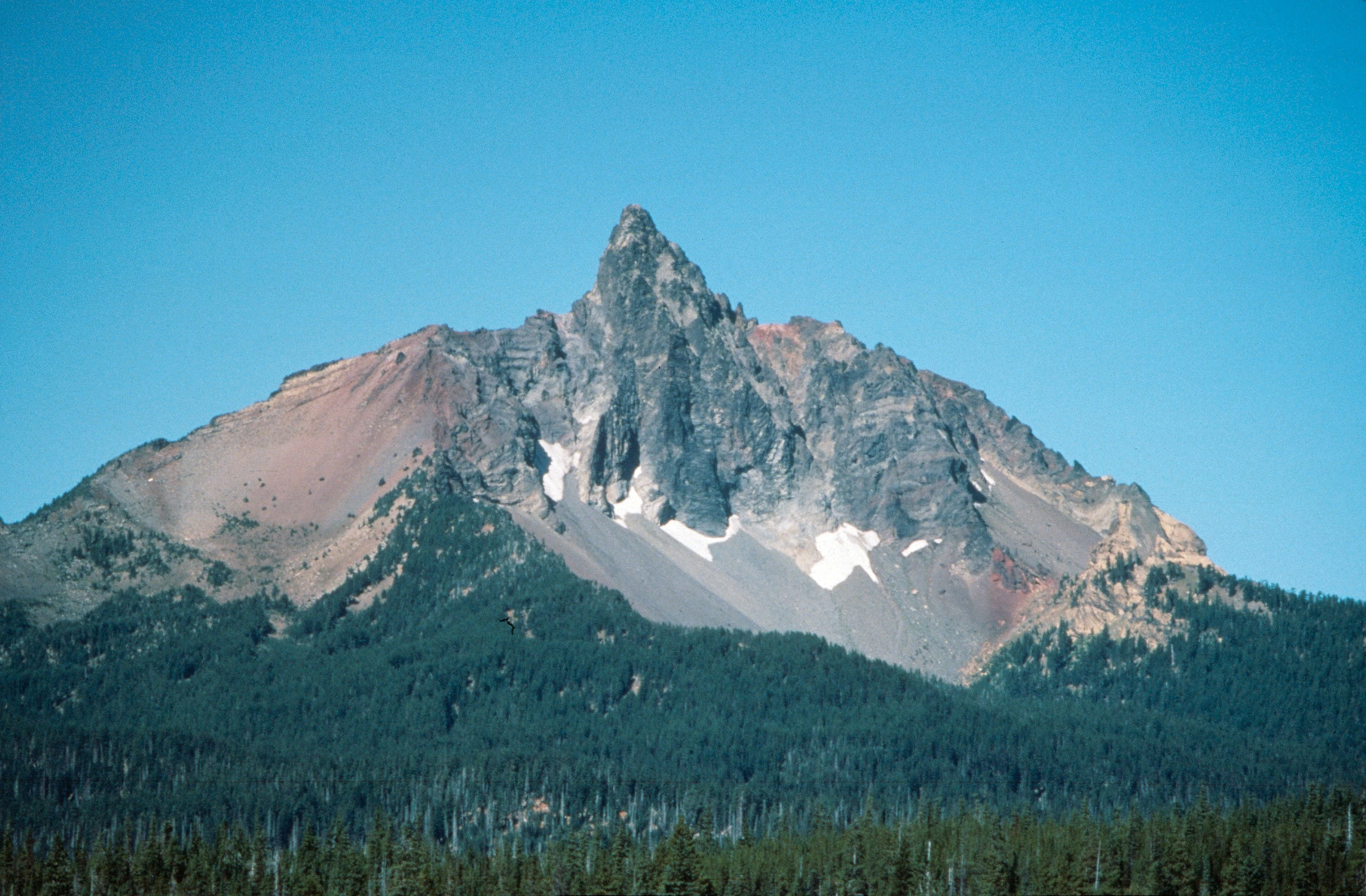A wide-angle, eye-level photograph of Mount Washington in Oregon against a clear blue sky, showing a jagged, rocky peak with patches of snow and a base covered in dense coniferous forest.