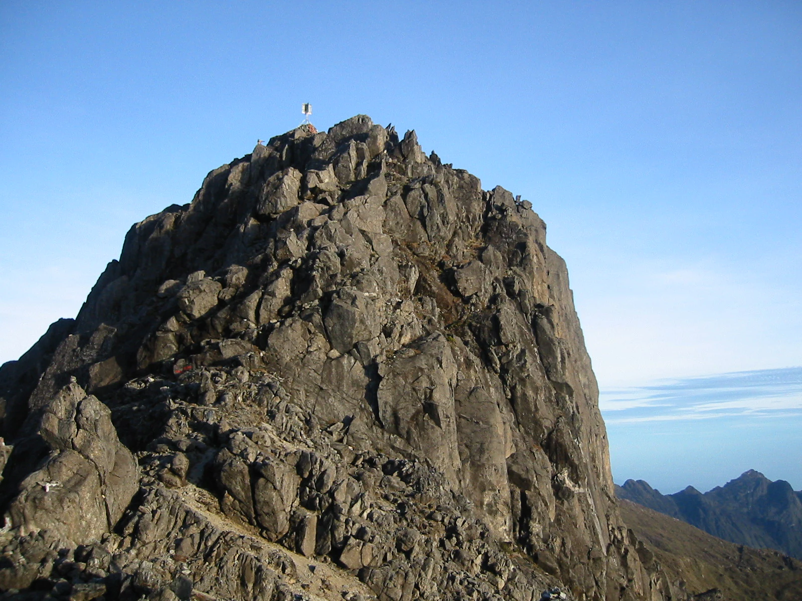 Rocky mountain peak with a small structure visible at the summit against a clear blue sky