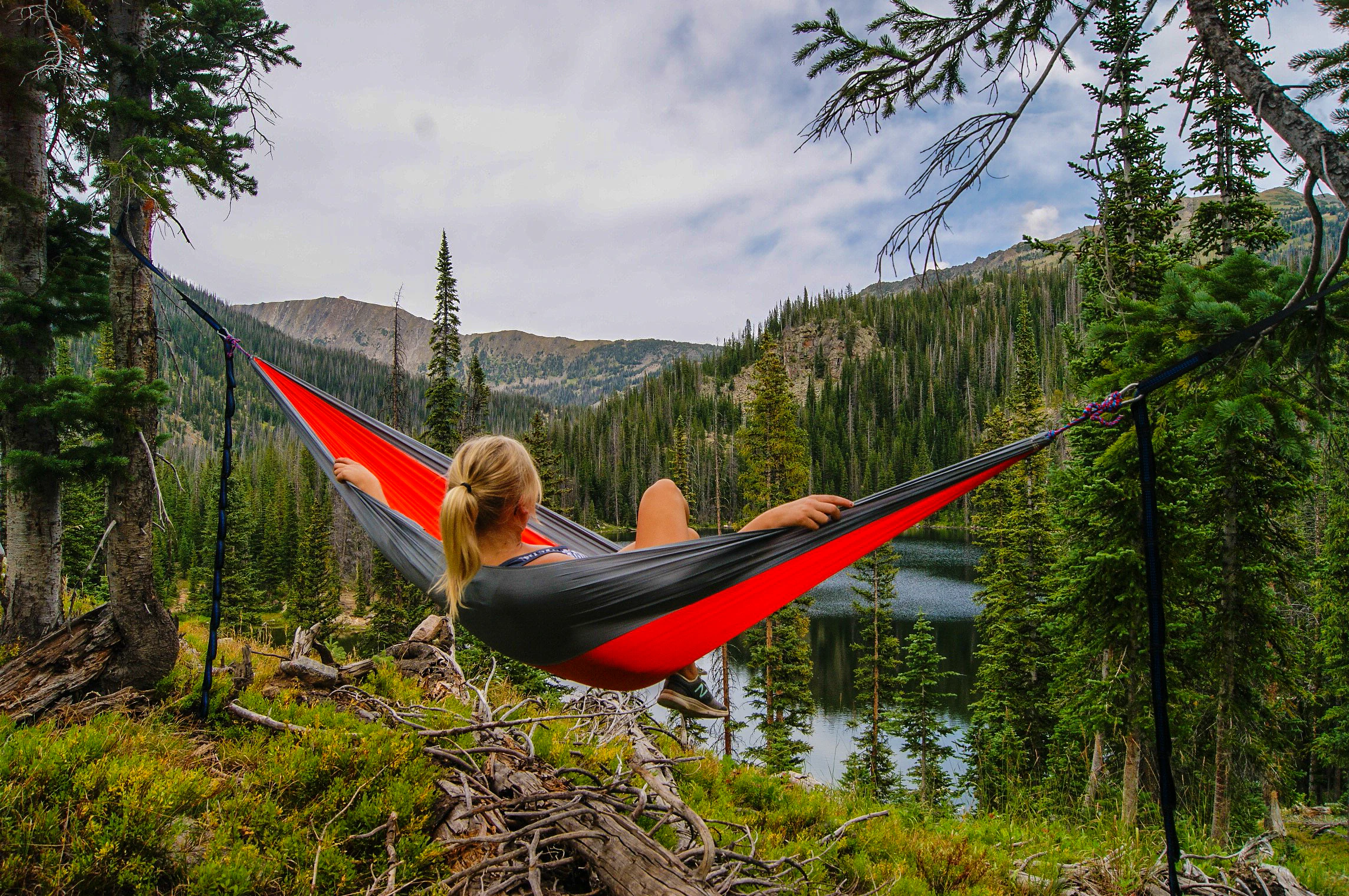 A person viewed from behind reclines in a bright red and grey hammock strung between evergreen trees, overlooking a small alpine lake and forested mountainside.