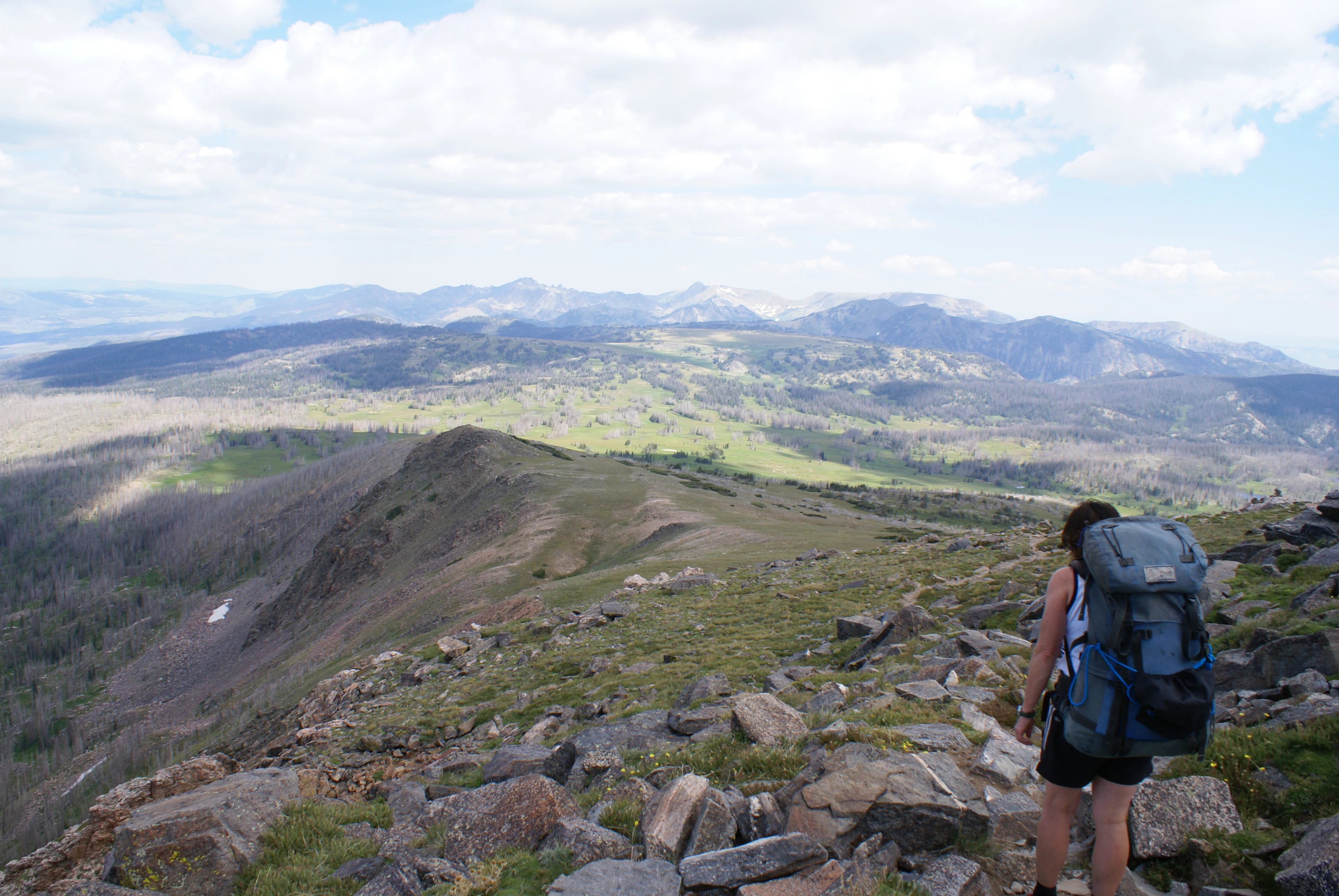 A hiker carrying a large blue backpack stands on a rocky, high-altitude trail overlooking a vast mountainous landscape under a cloudy sky.